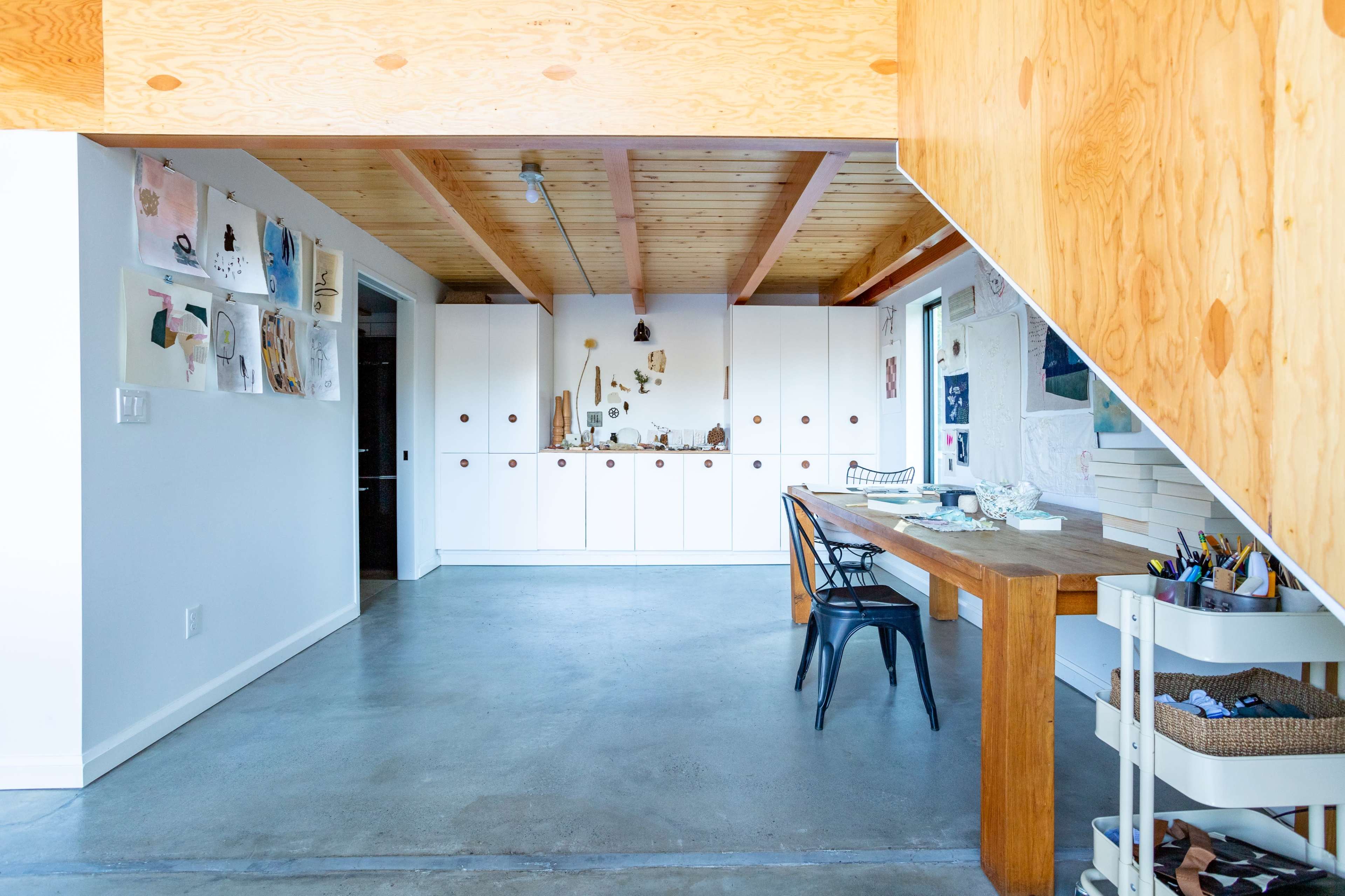 A modern interior space with a wooden table and chairs, concrete flooring, and a wall adorned with various artworks and storage cabinets.