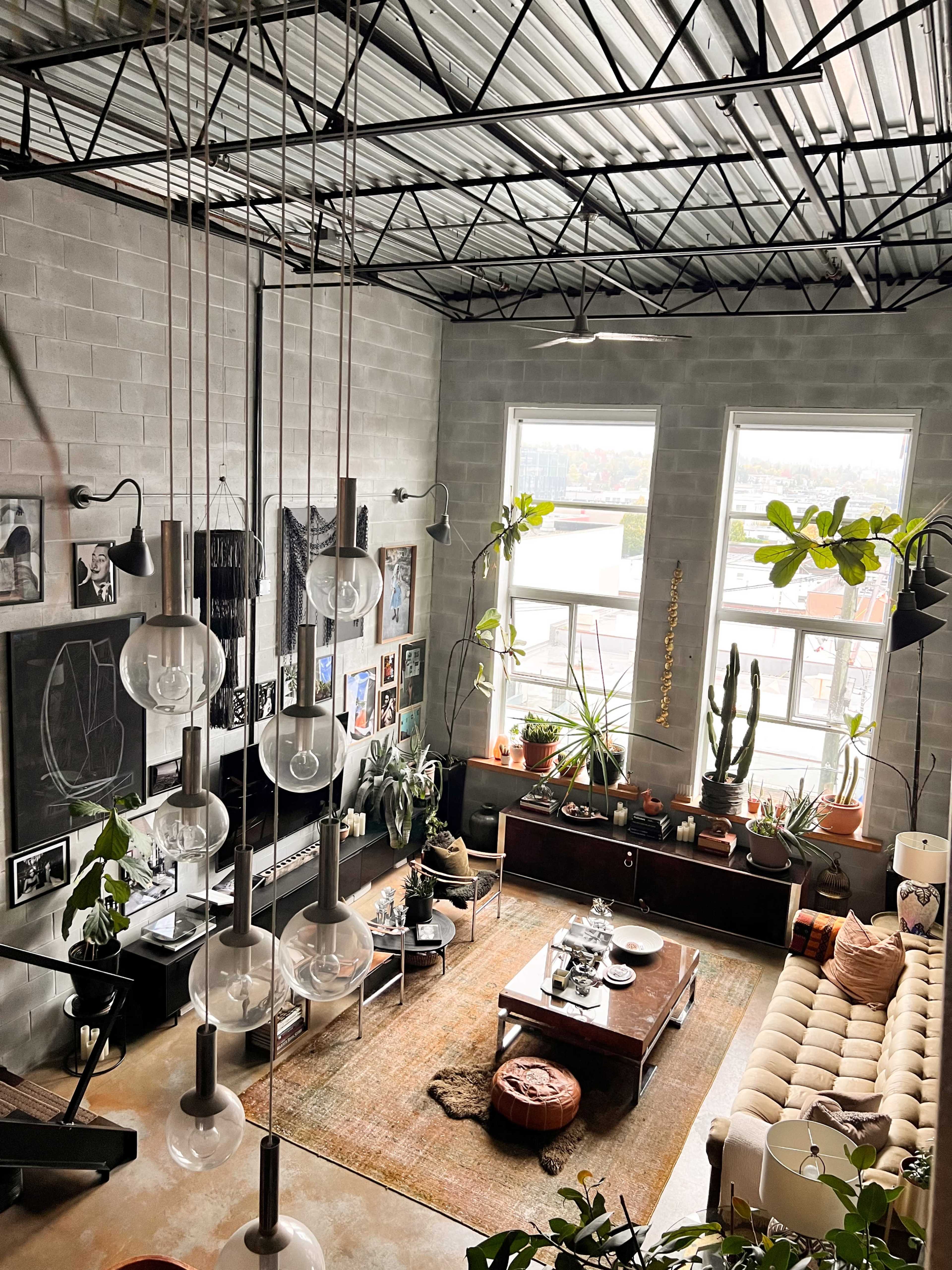A spacious, industrial-style living room with large windows, a variety of indoor plants, and a mix of modern and vintage furniture arranged around a central coffee table.