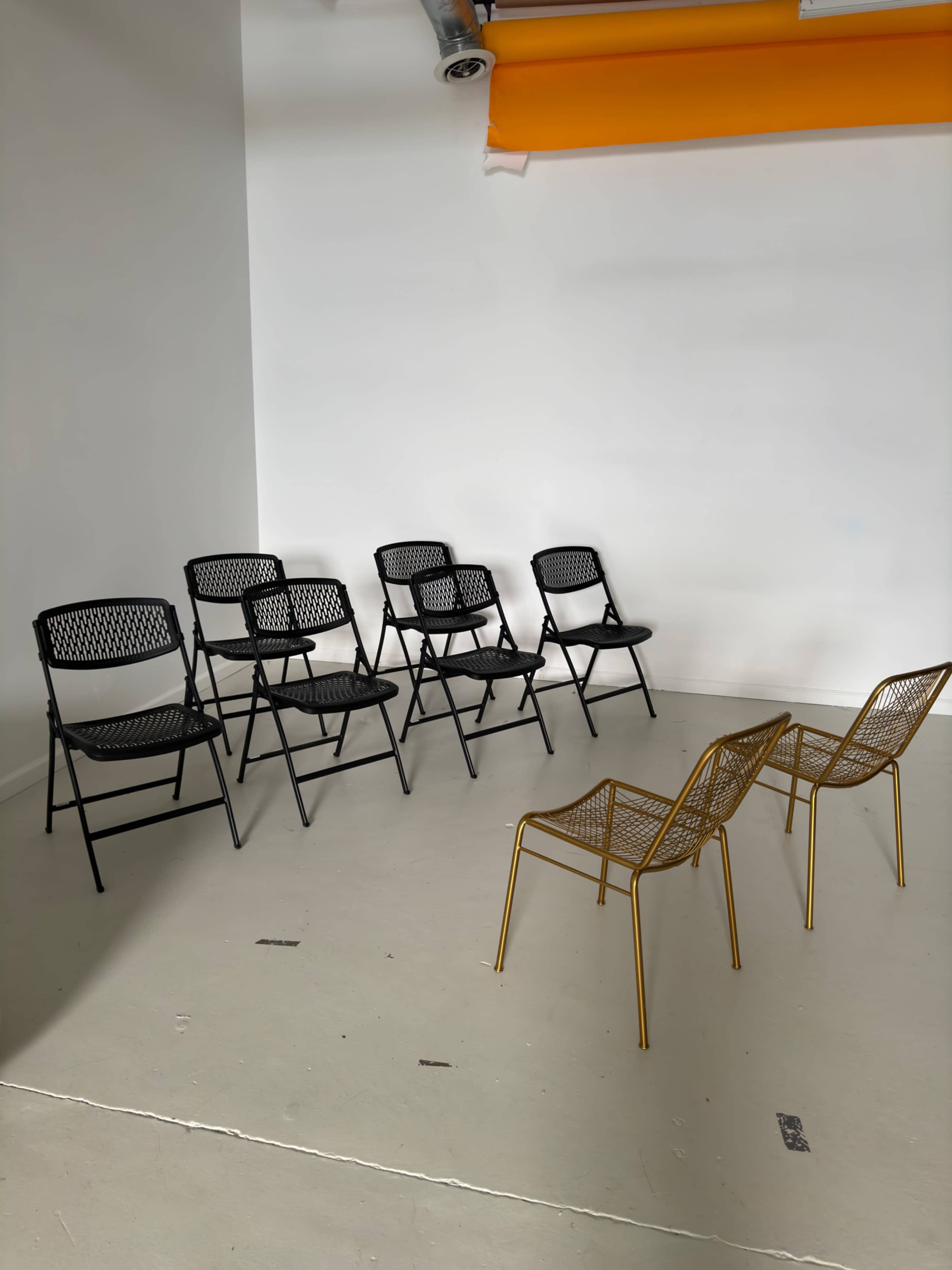 A minimalist space with six black folding chairs lined up against a white wall, and two gold wire chairs facing them.