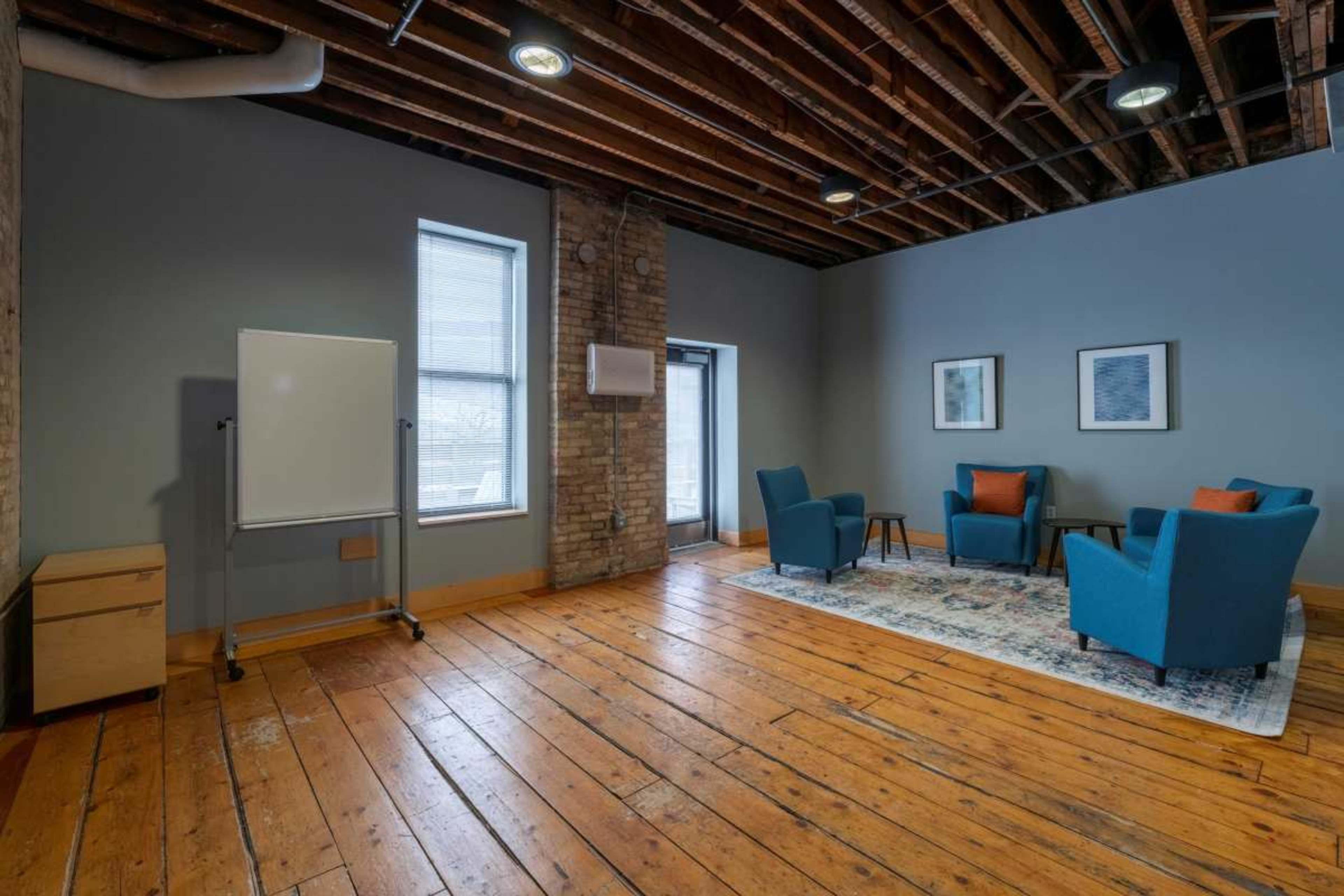 The image shows a spacious room with wooden floors, featuring two blue armchairs, a small table, a whiteboard on a stand, and windows that allow natural light in.