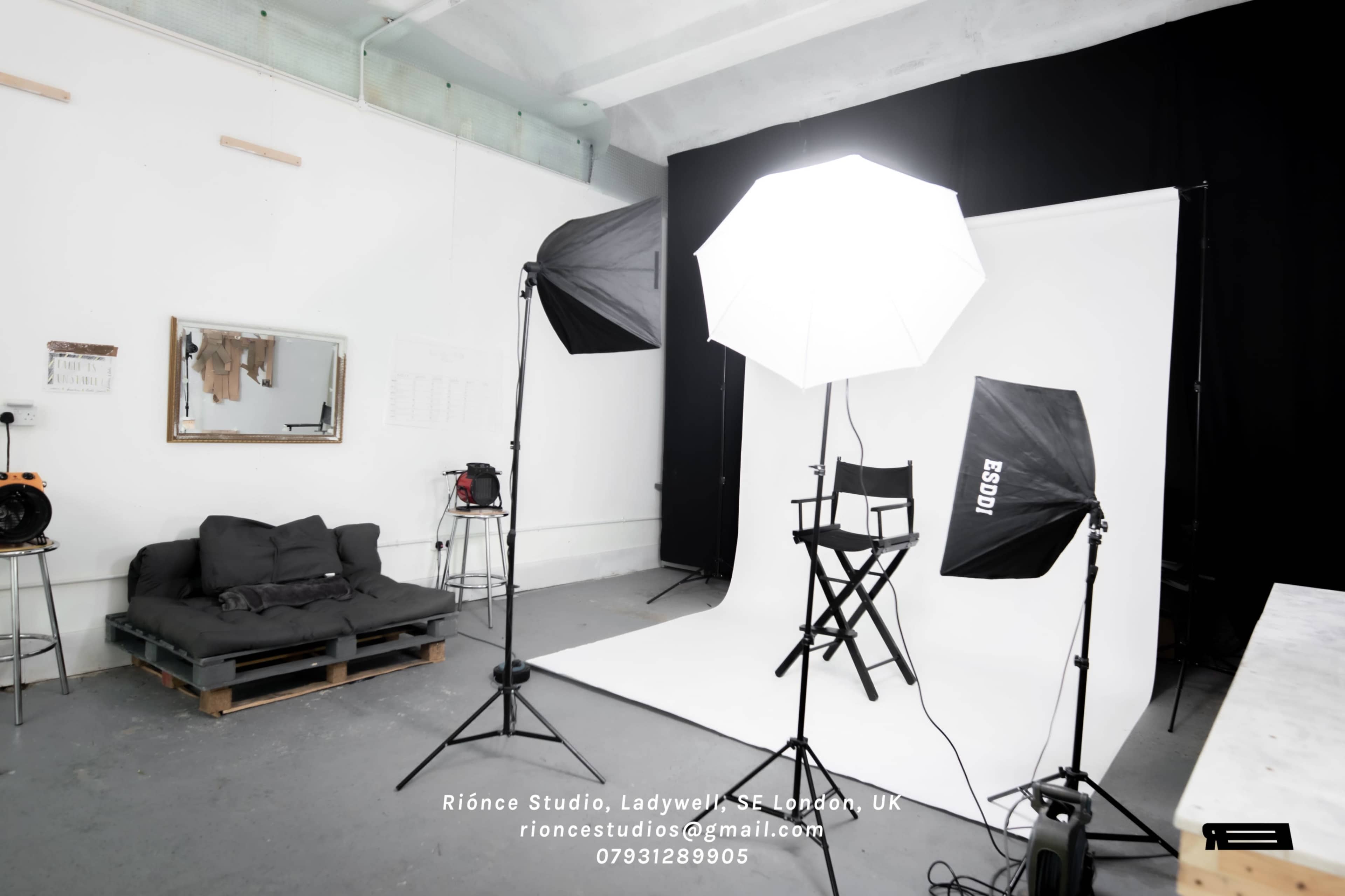 A photography studio setup featuring a white backdrop, lighting equipment, and a director's chair, with a couch in the corner.