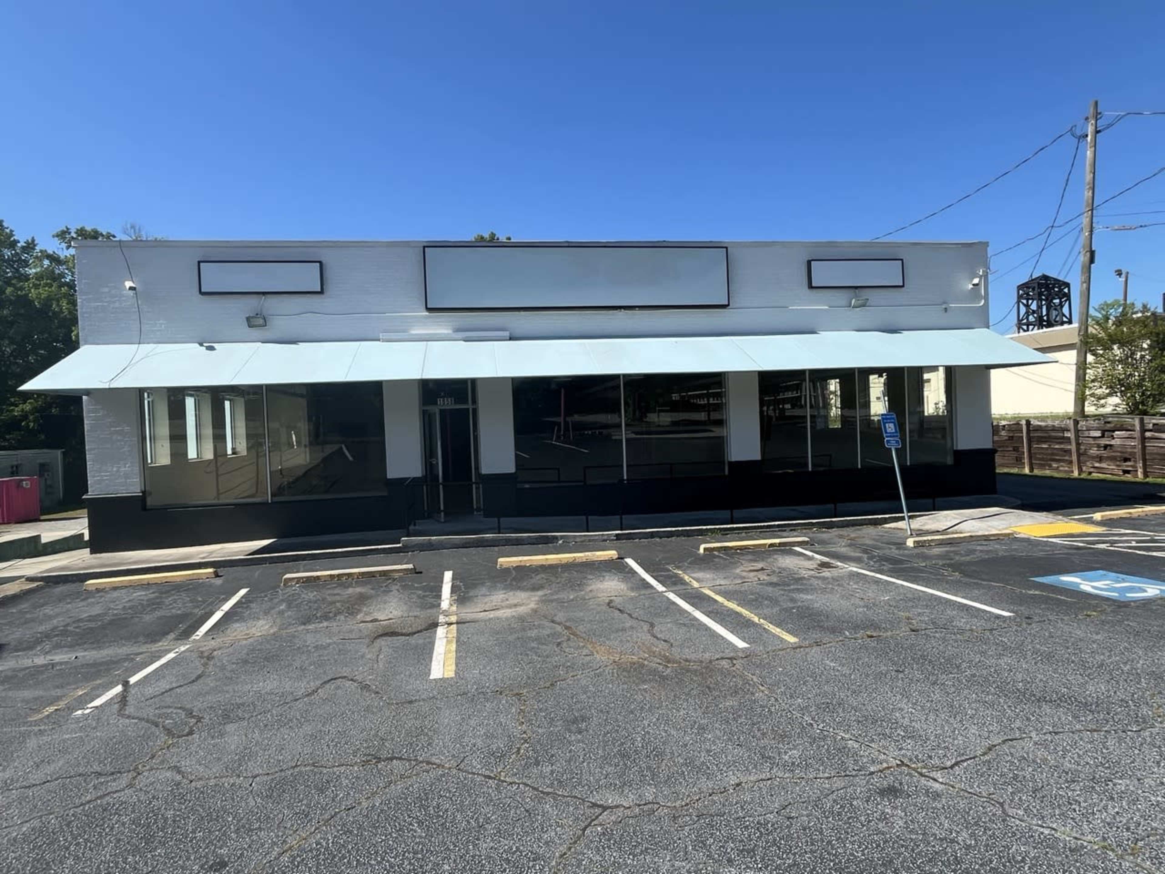 A vacant commercial building with a large awning and multiple windows sits in an empty parking lot under a clear blue sky.