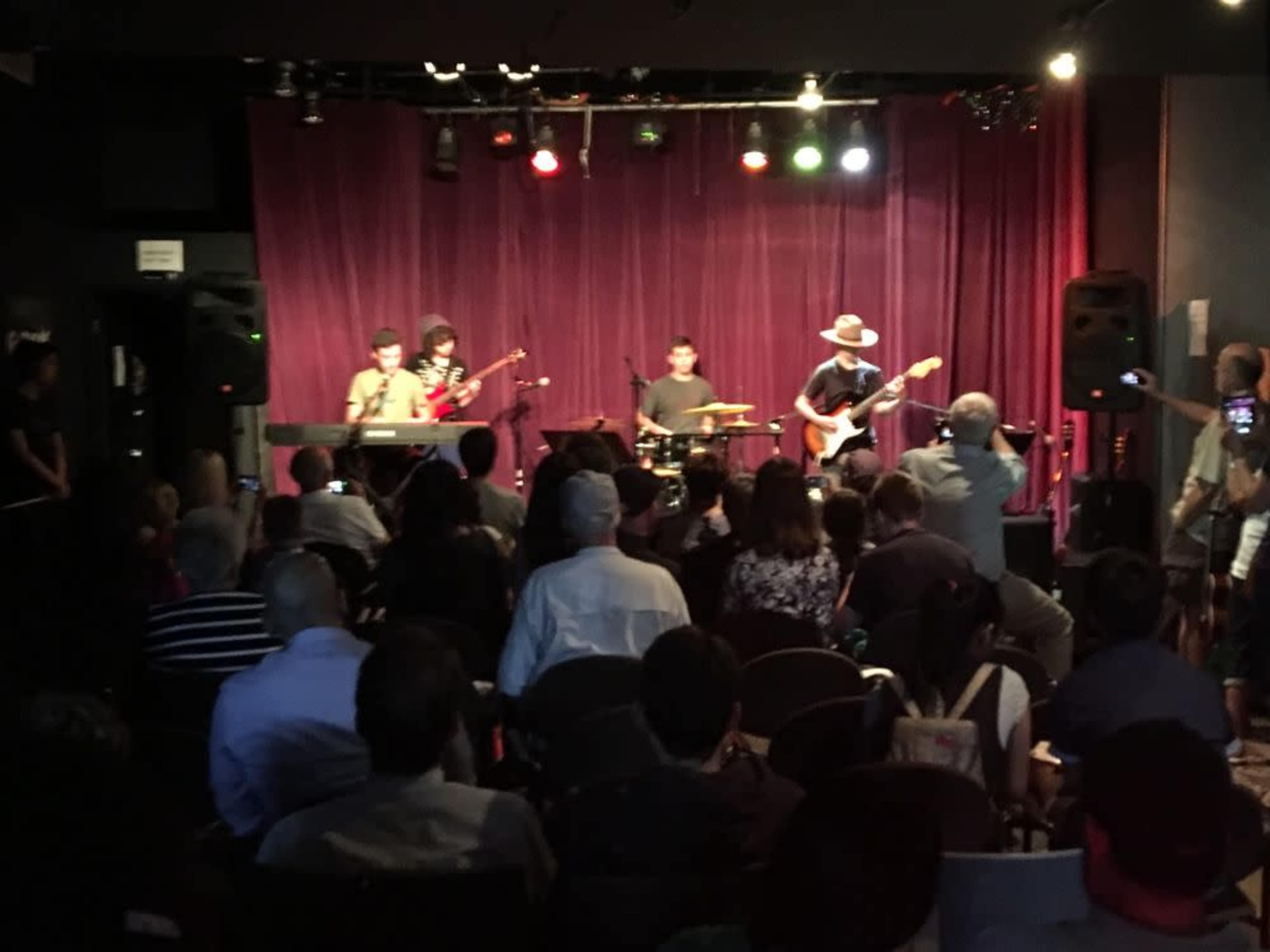 A band performs on stage in front of an audience in a dimly lit venue with purple drapes.