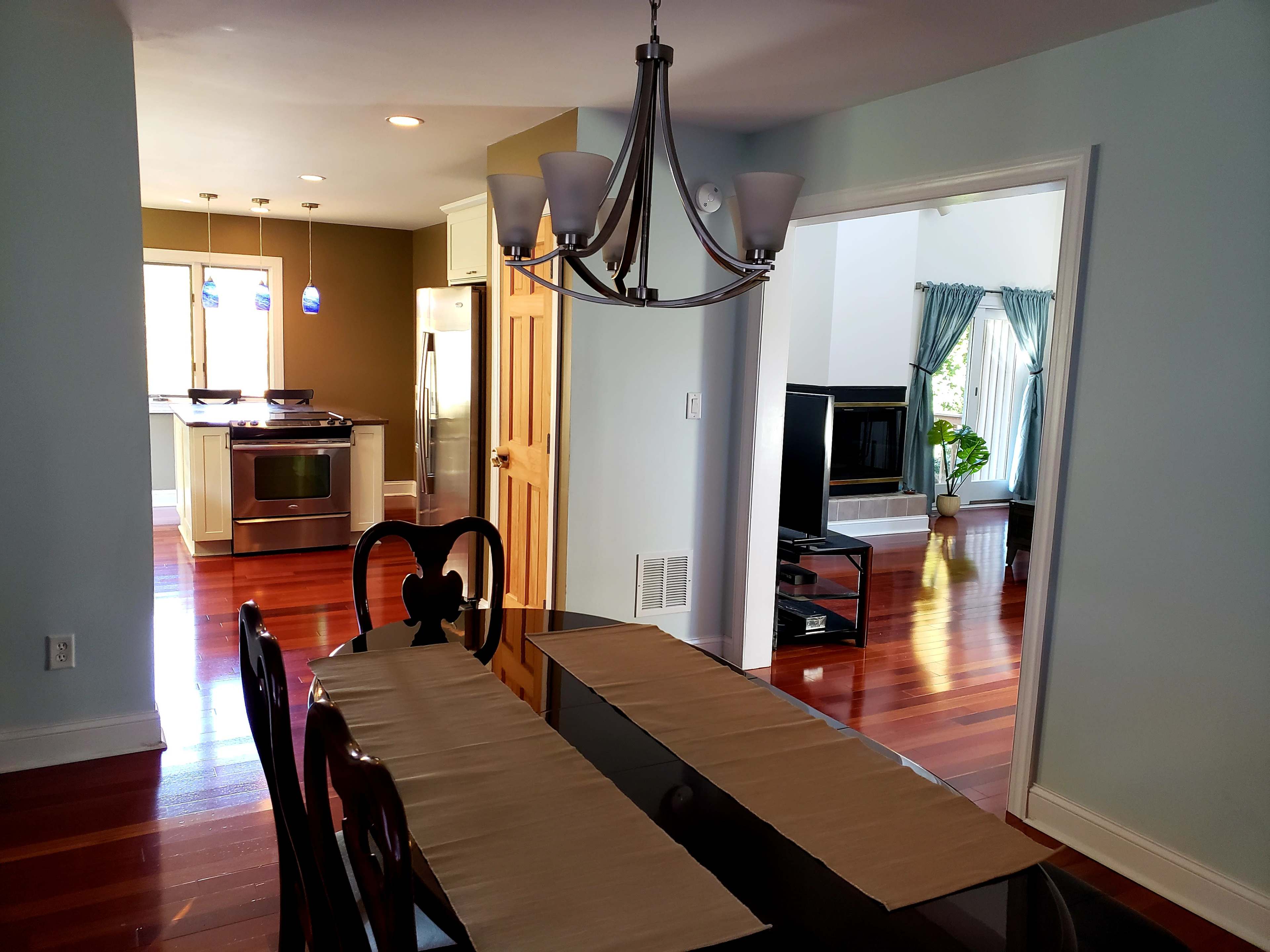 A dining area with a table set for meals, leading into an open kitchen and a living room with large windows and a fireplace.