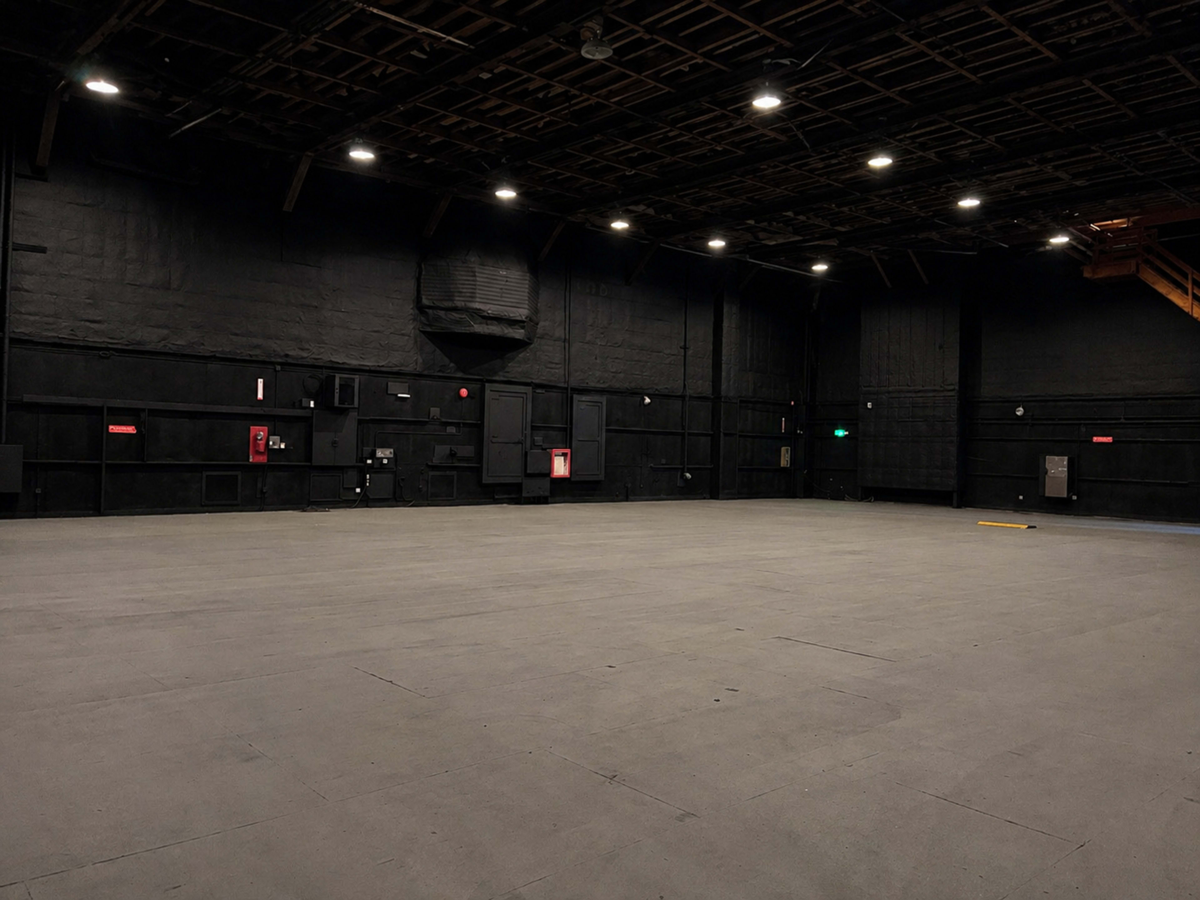 The image shows a large, empty room with dark walls and a flat, spacious floor, illuminated by overhead lights.