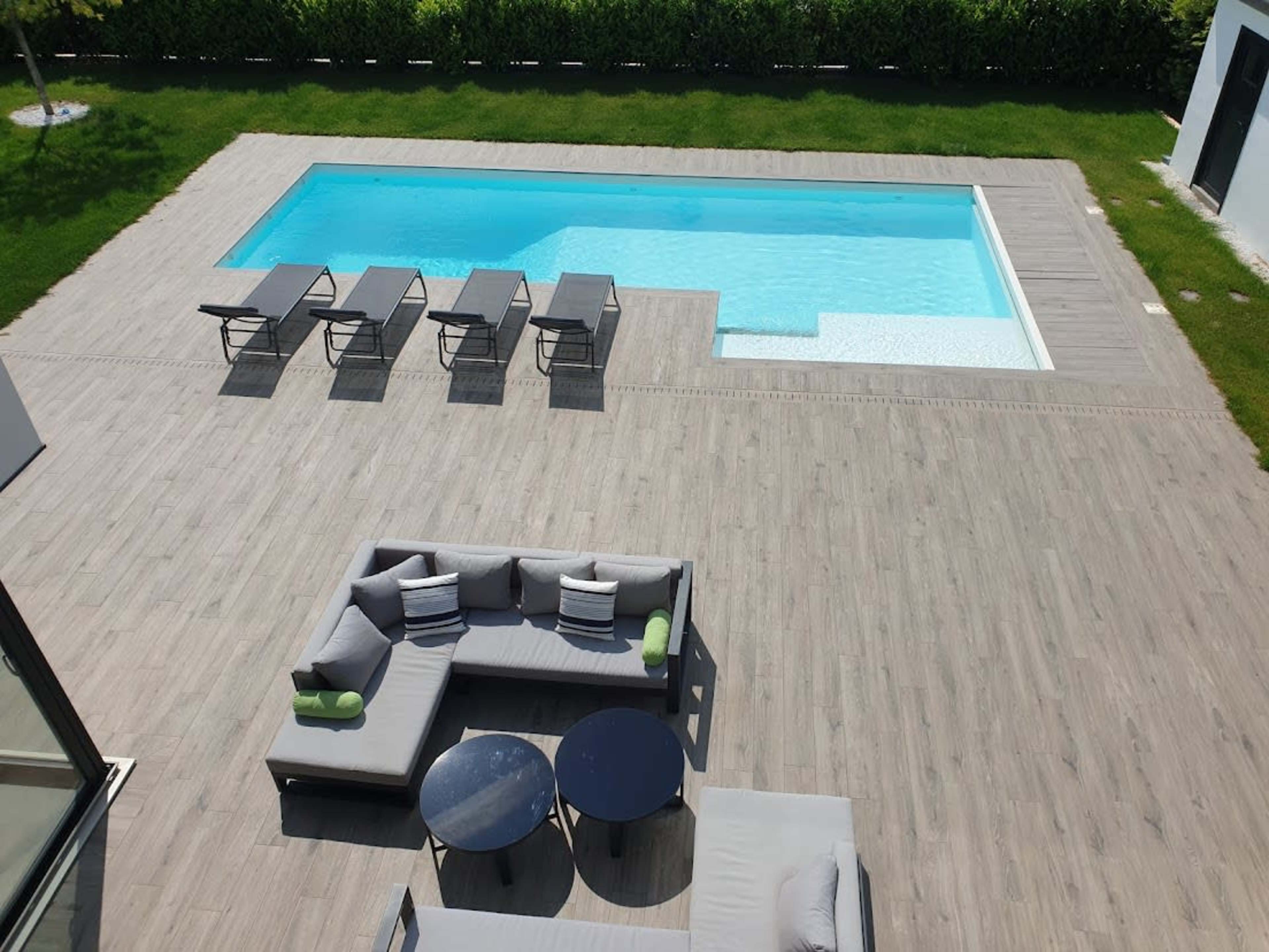 A modern outdoor space features a rectangular pool surrounded by wooden decking, lounge chairs, and a seating area with gray couches and black tables.