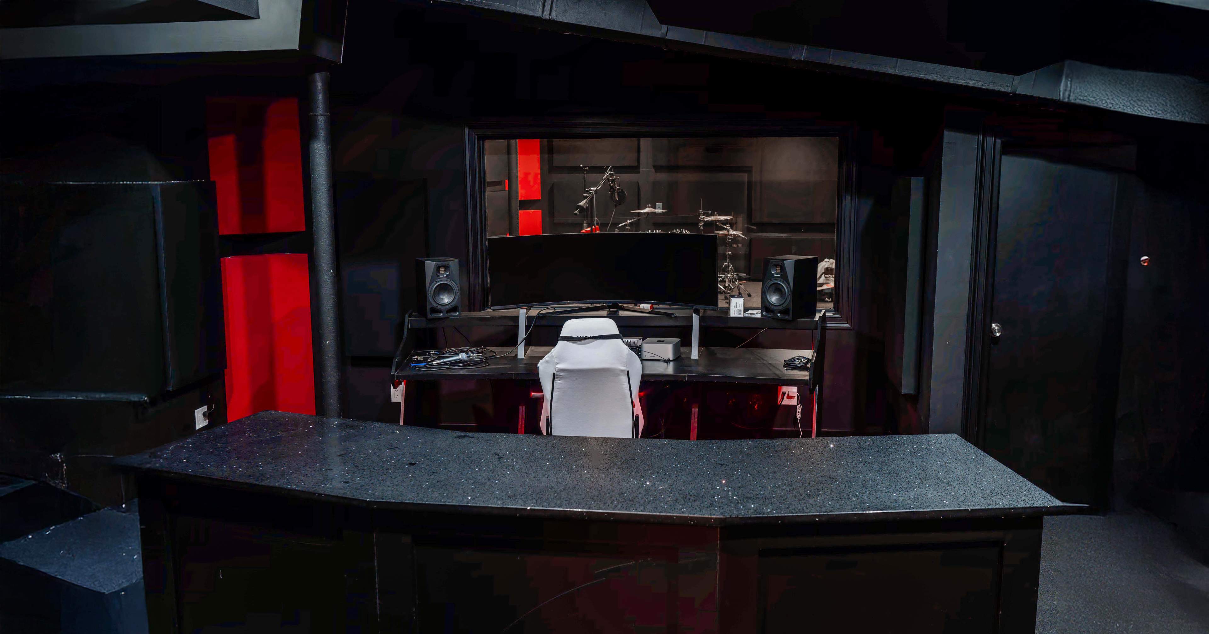 The image shows a music studio with a central desk, a computer monitor, audio speakers, and a view into a drum area, all set against a dark, red-tinged backdrop.