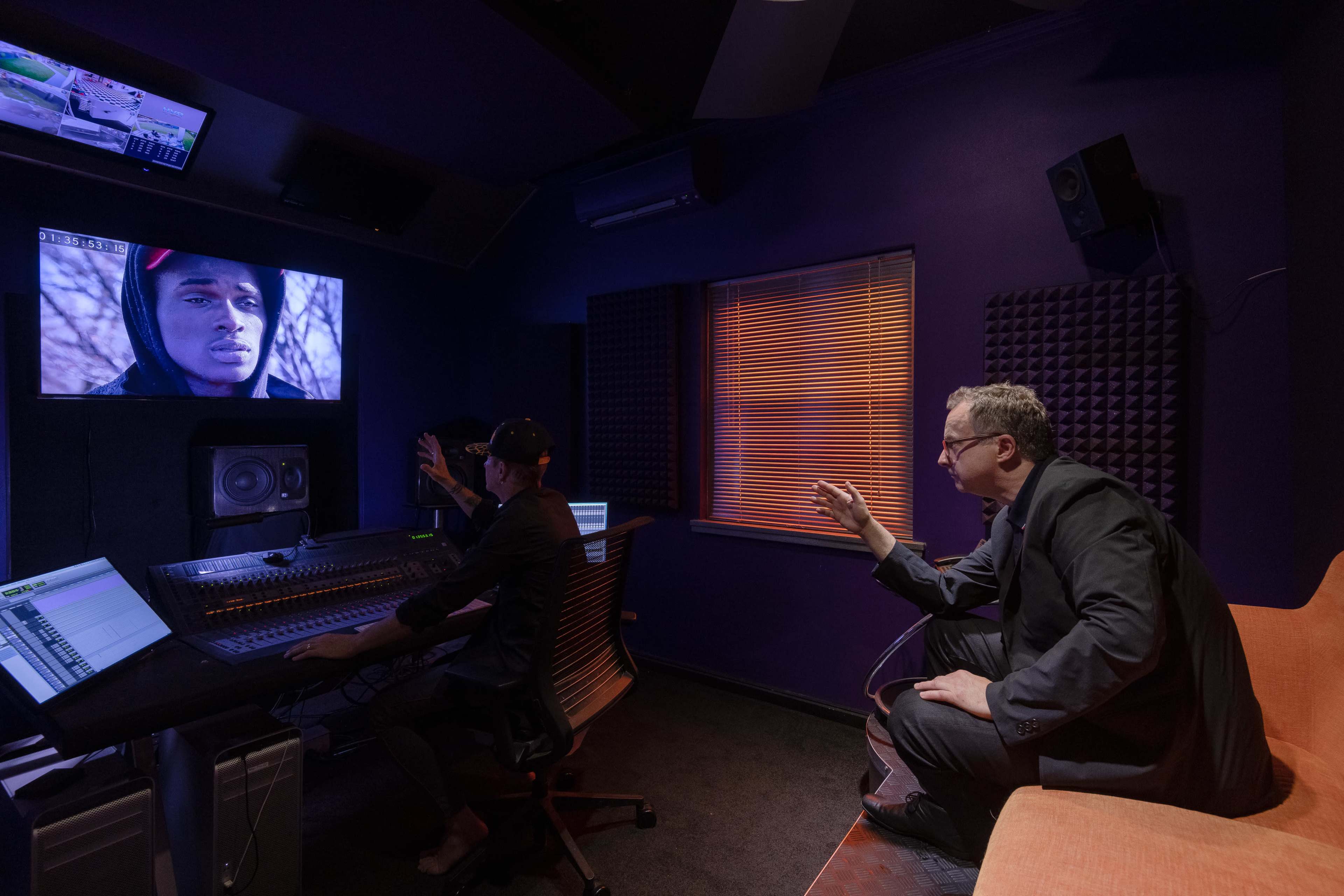 A man in a suit sits on an orange chair while another person works at a mixing console in a recording studio, with a large screen displaying a close-up of a young man.