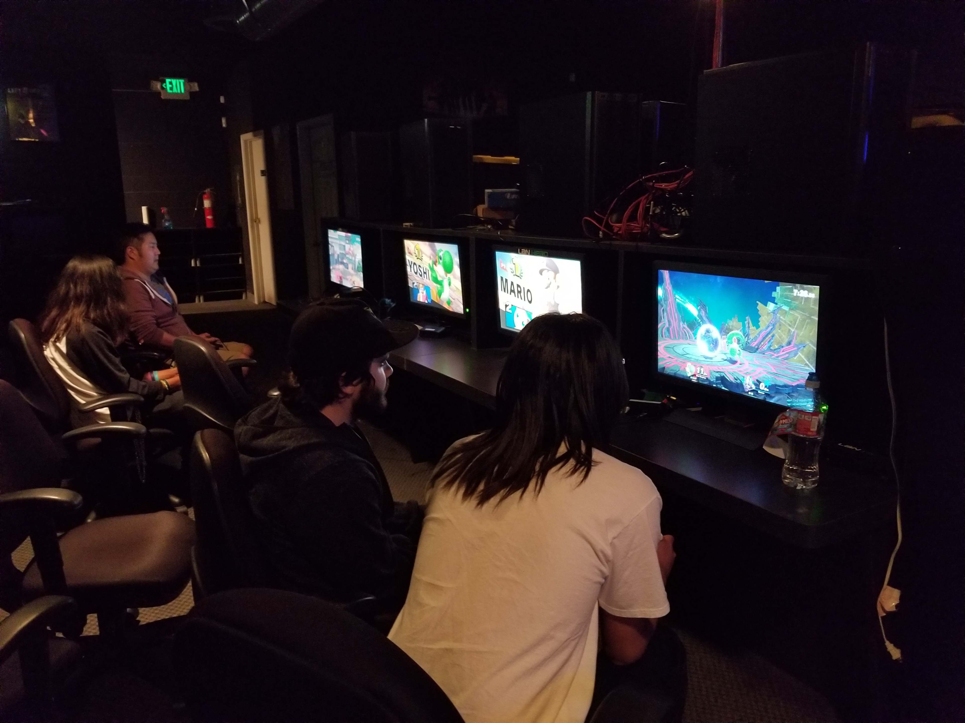 A group of four people are seated at gaming stations, focused on different video games displayed on screens in a dimly lit room.