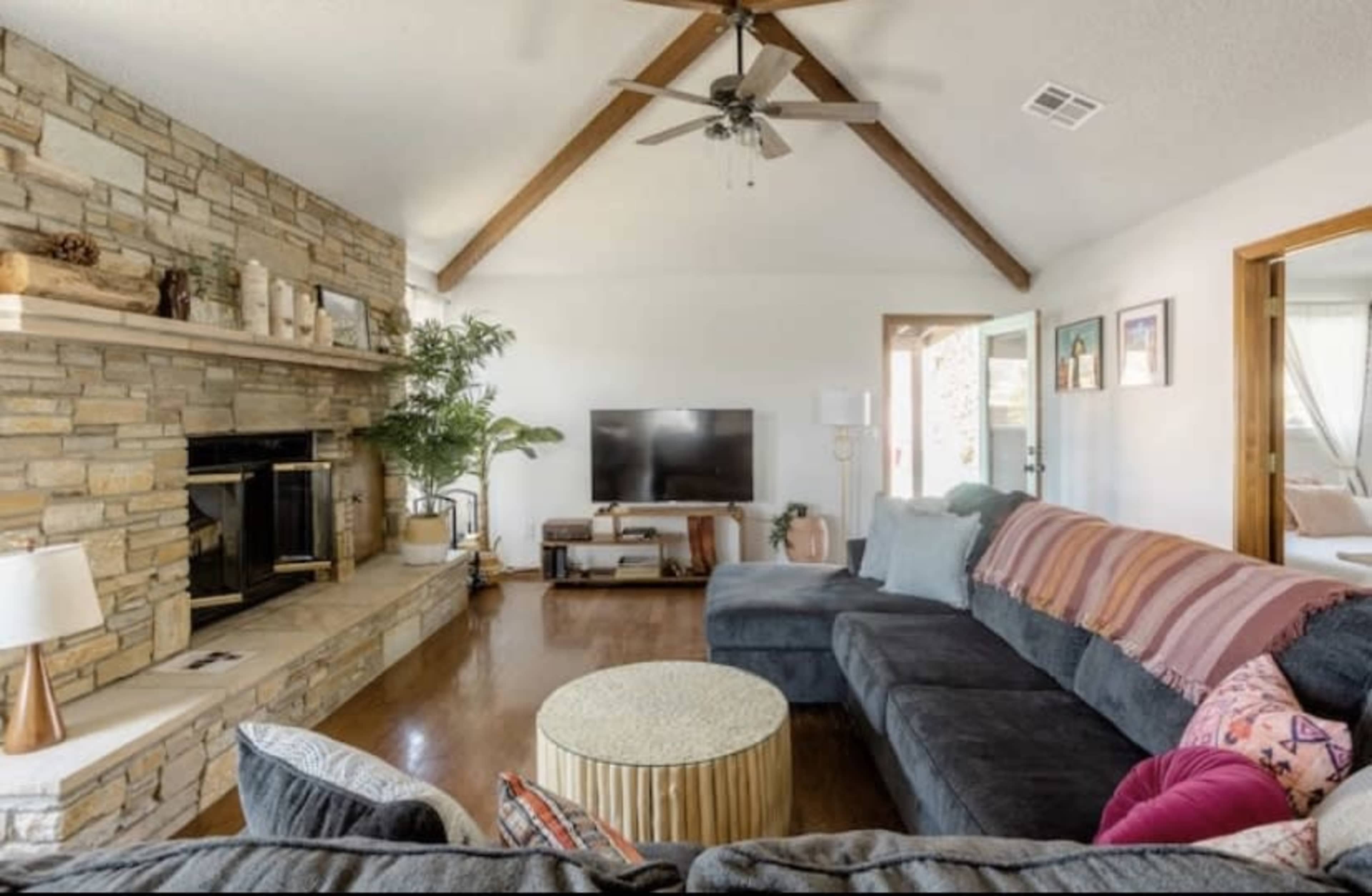 The living room features a stone fireplace, a large sectional sofa with colorful throw pillows, and a round coffee table, all set under exposed wooden beams.