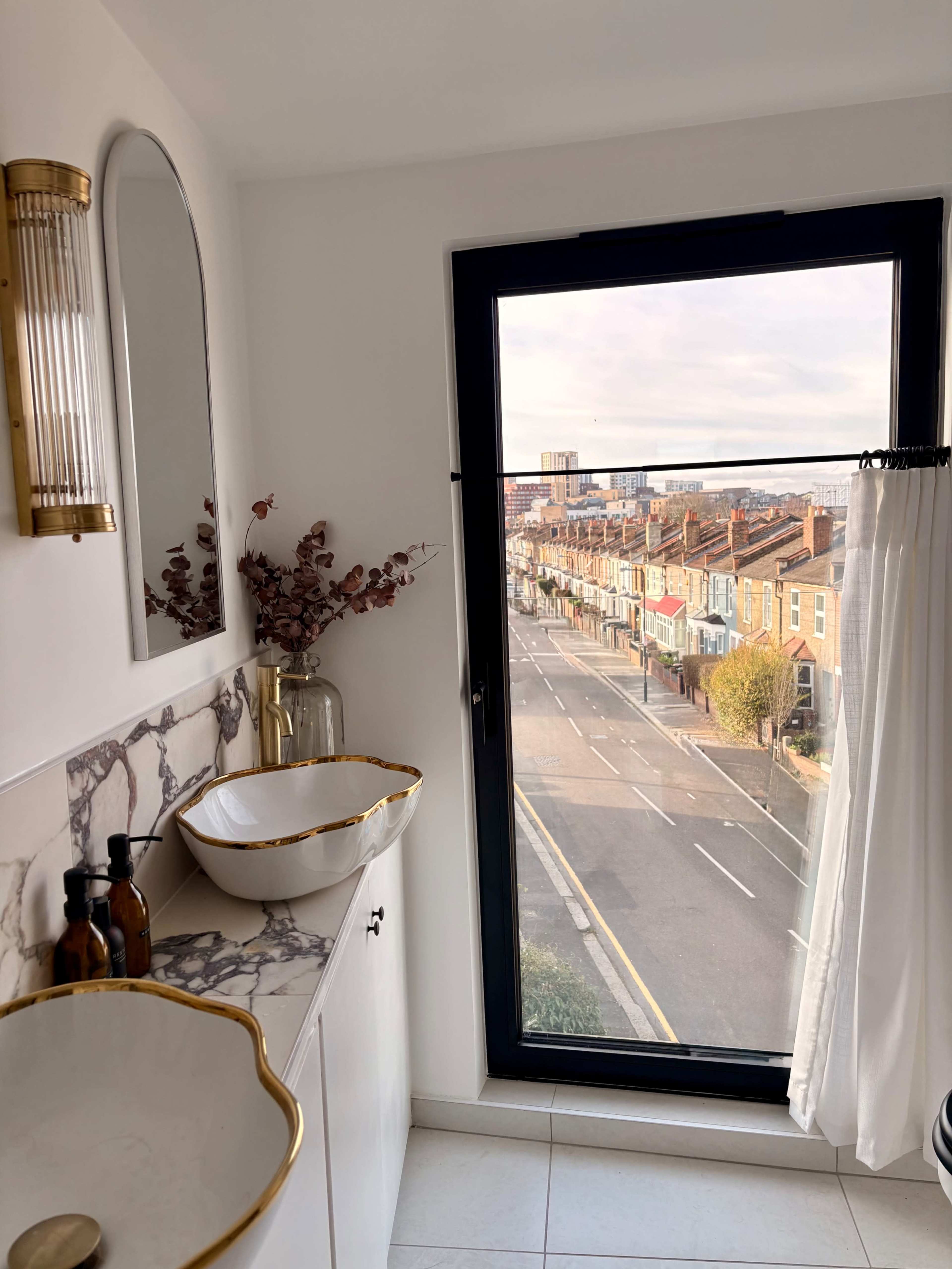 The image shows a modern bathroom with a marble countertop, two sinks, and a large window overlooking a street lined with houses.