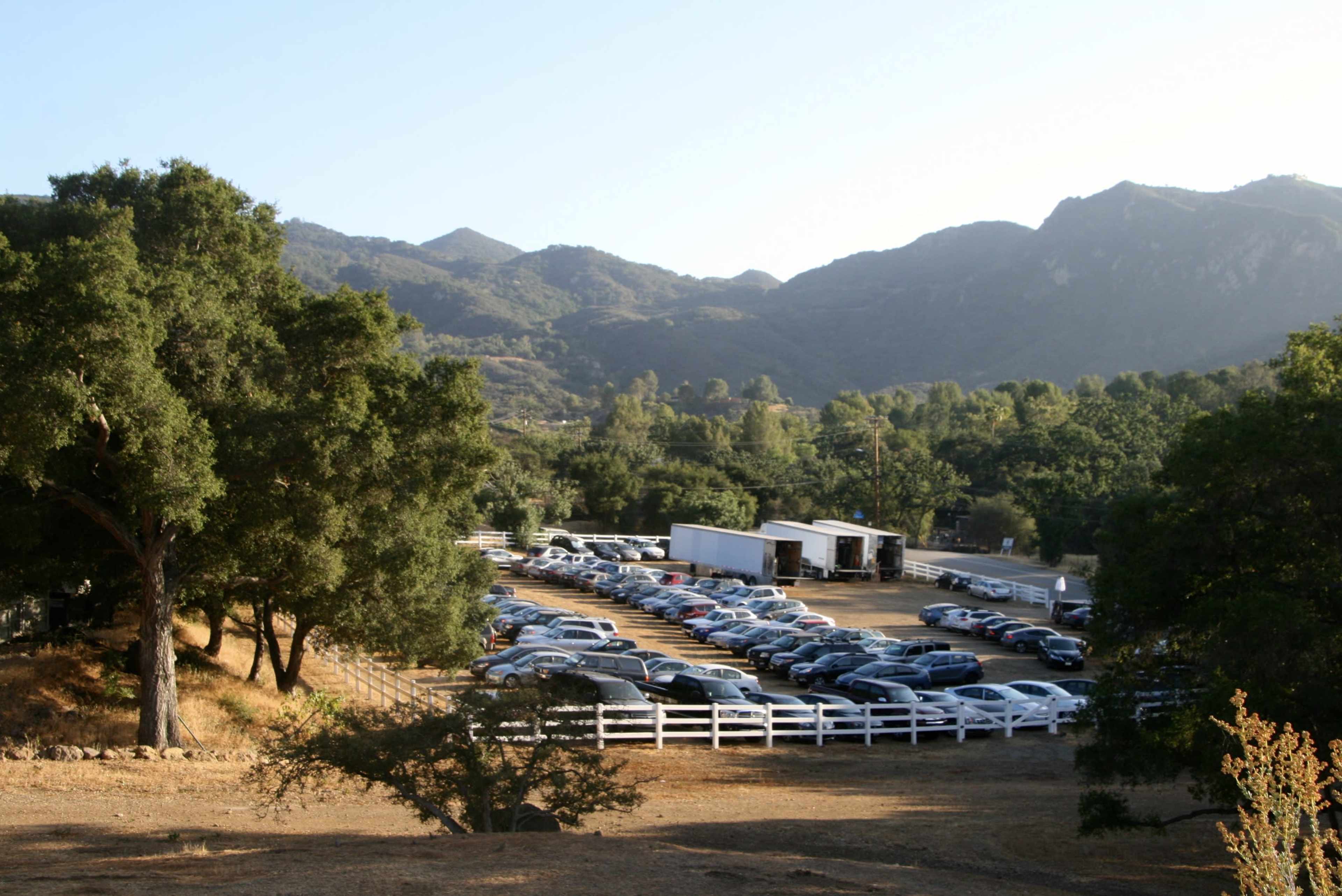 A large parking lot filled with cars is situated in a rural landscape surrounded by trees and mountains.
