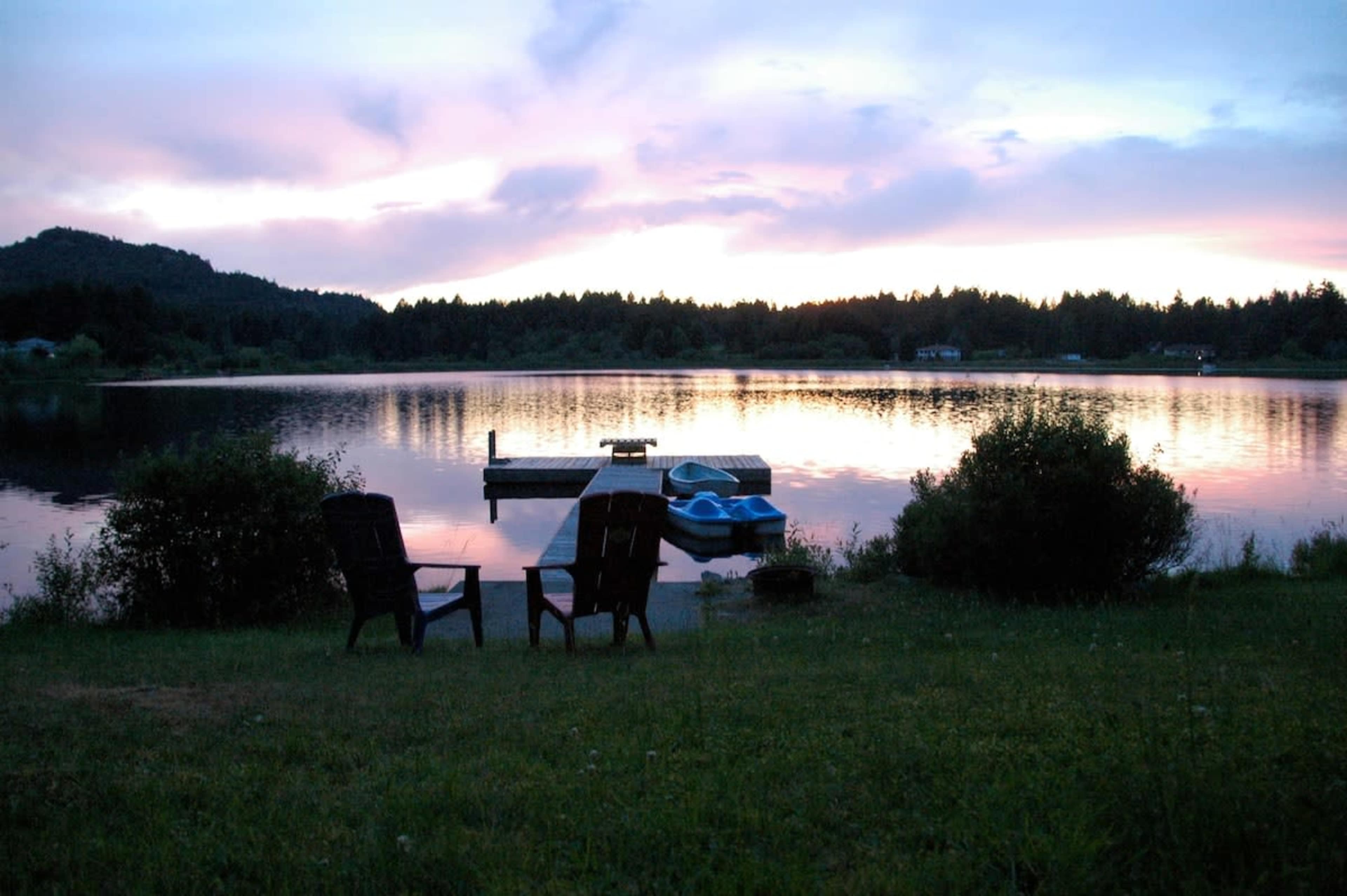 Two chairs sit on the grass by a calm lake, with a dock and a blue boat visible in the water beneath a colorful sky at sunset.