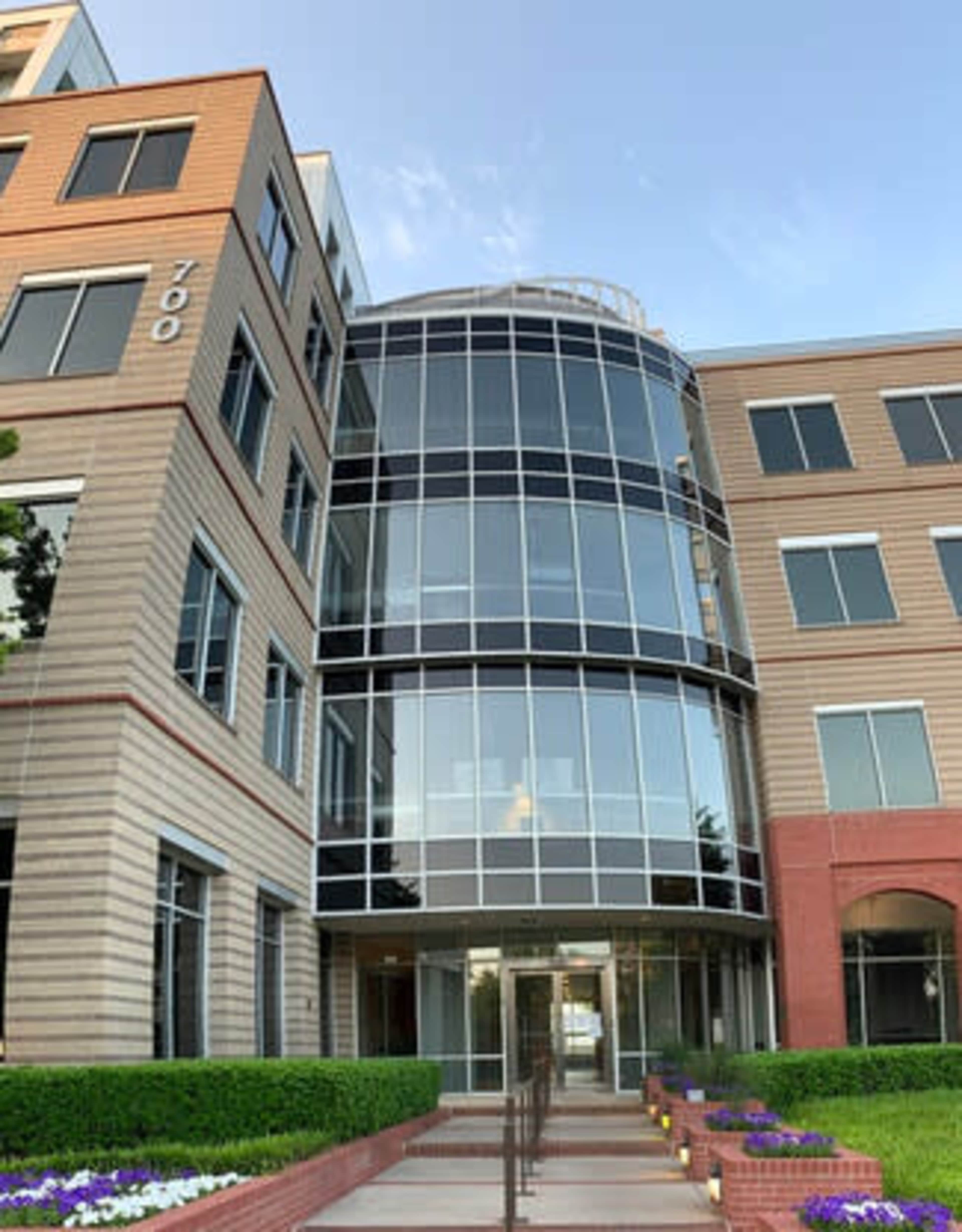 The image shows a modern office building with large glass windows and a landscaped entrance featuring a pathway and flower beds.