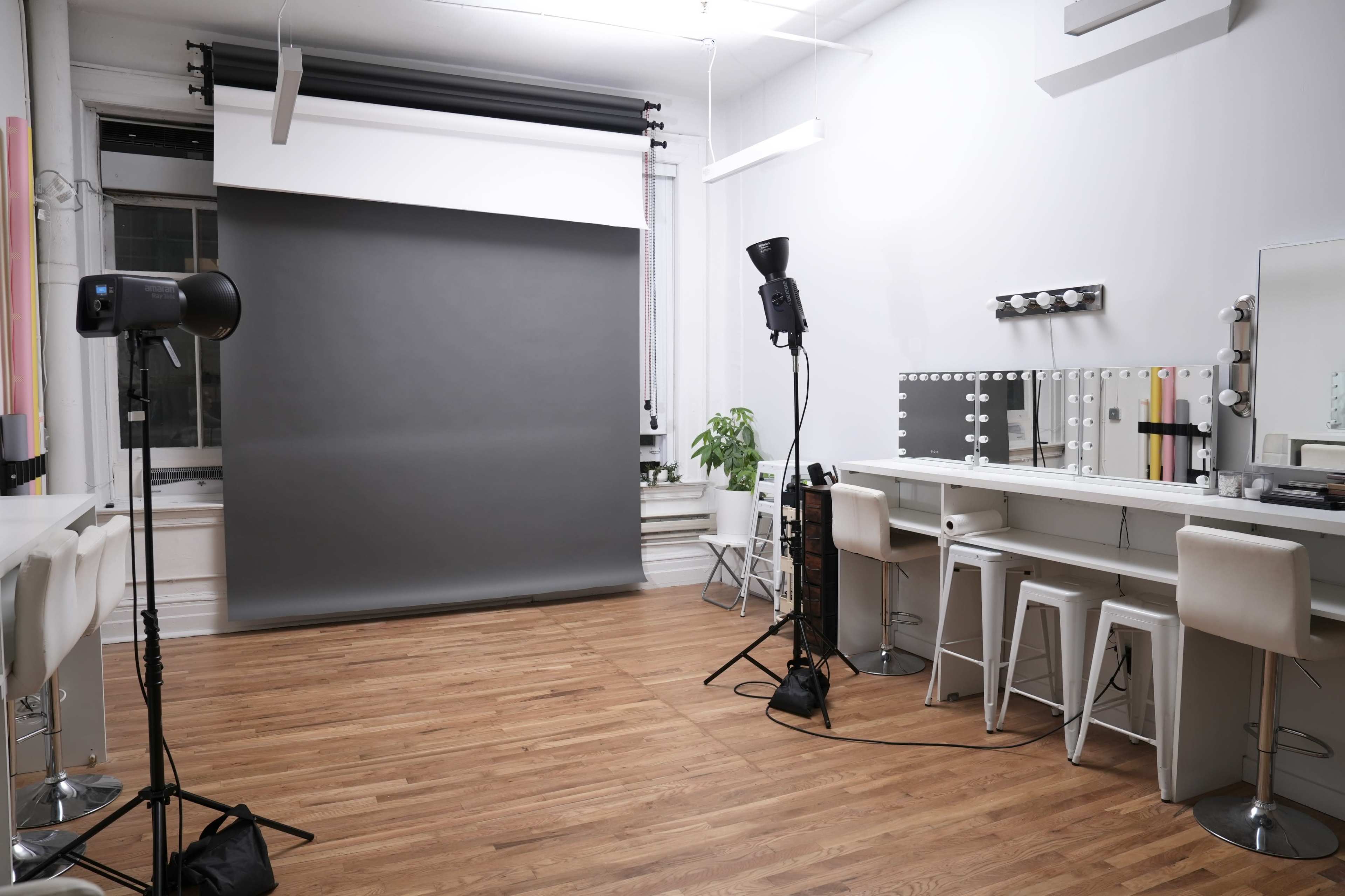 The image shows a well-lit photography studio featuring a grey backdrop, makeup station with lights, and a wooden floor.