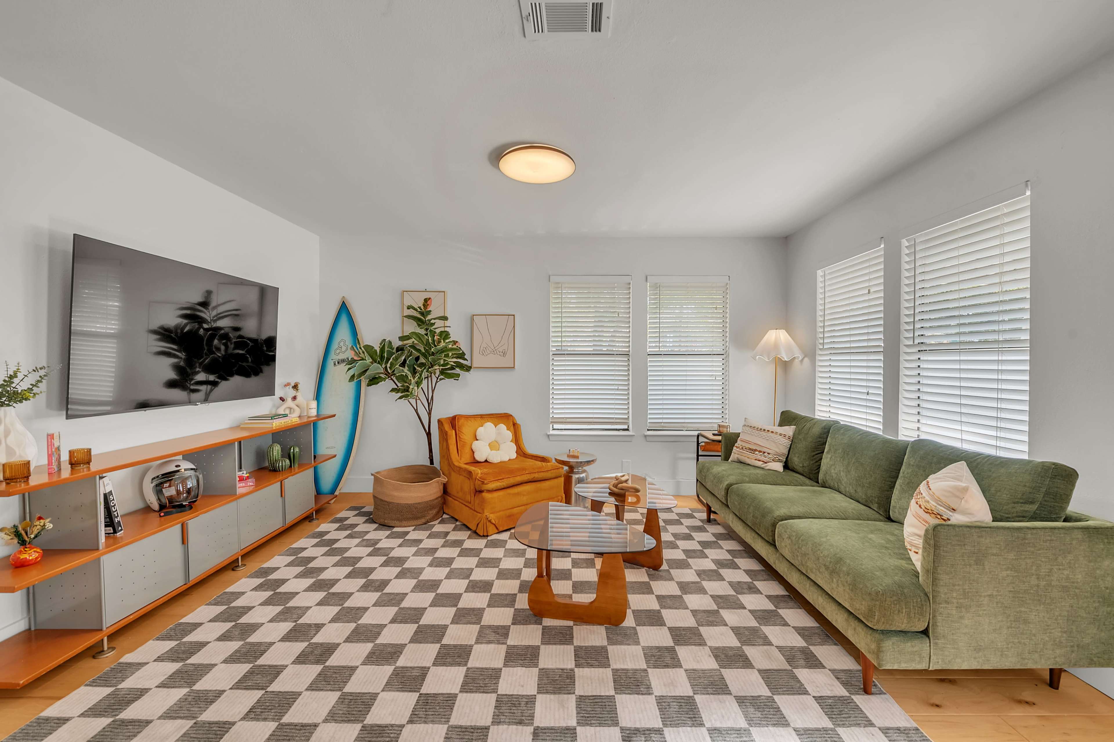 The living room features a green sofa, a mustard yellow armchair, a coffee table, and a large television against a white wall, with a checkered rug and house plants adding to the decor.
