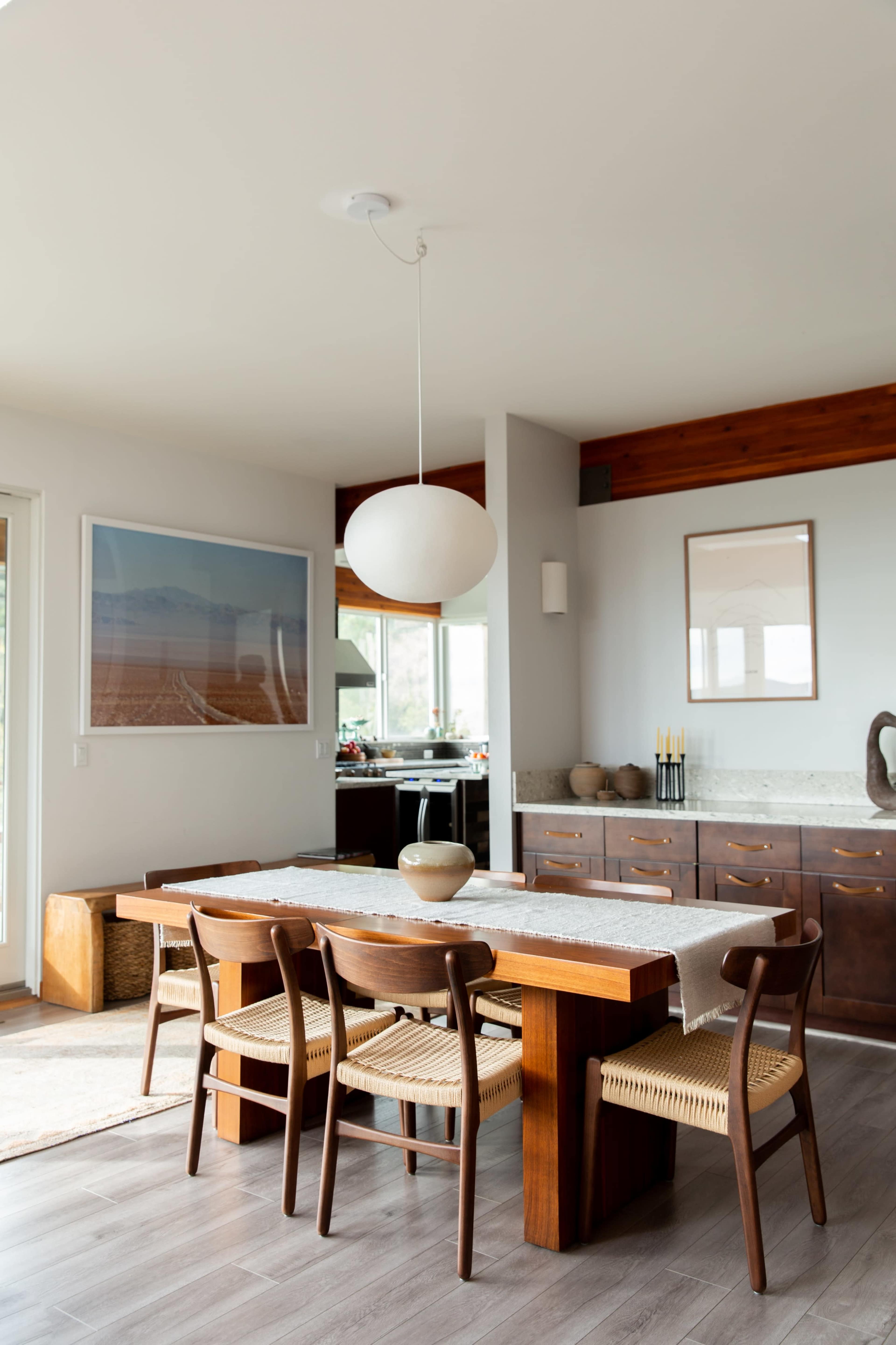 A dining area features a wooden table surrounded by chairs, with a large pendant light above and a sideboard against the wall.