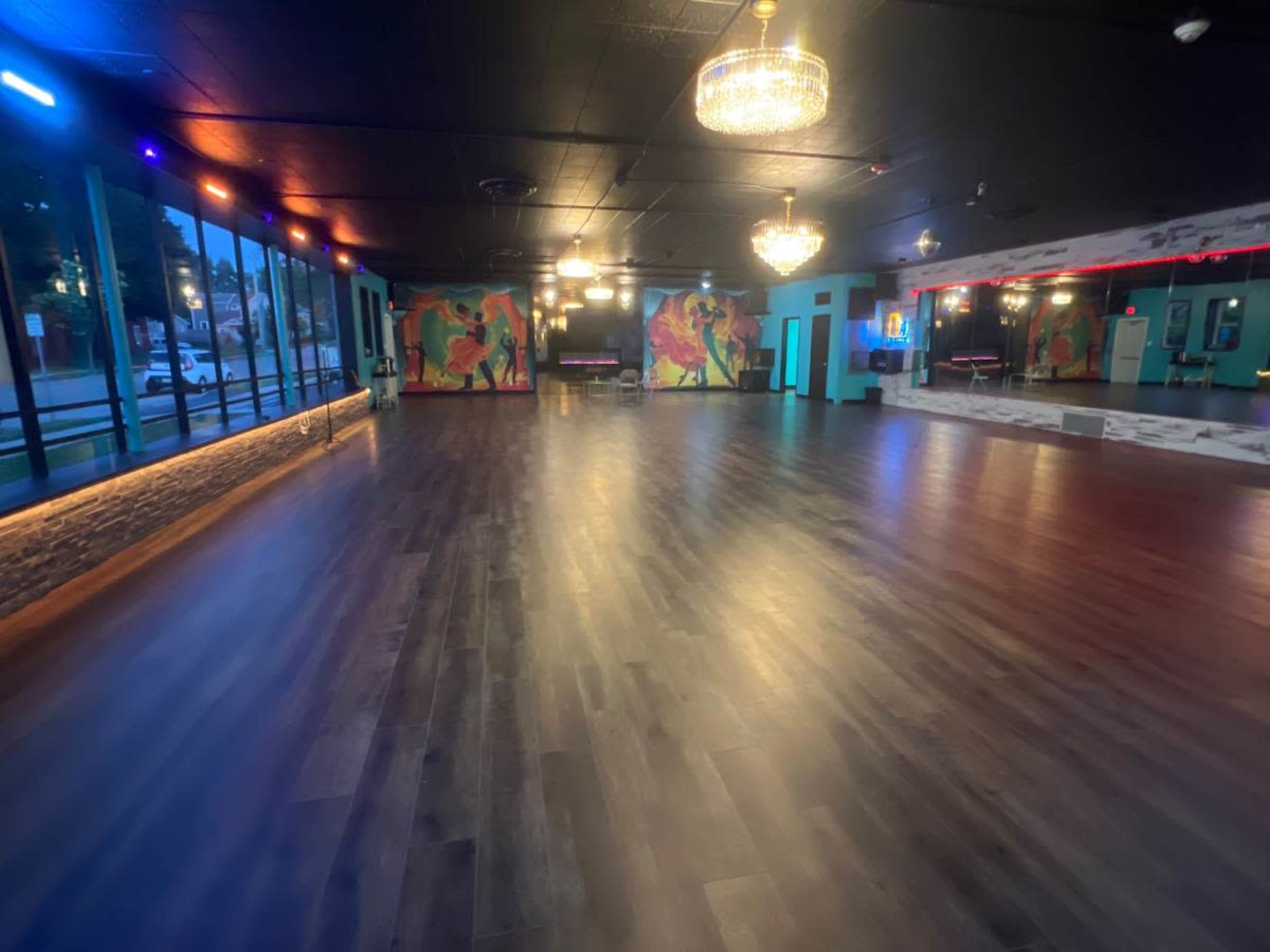 The image shows a dance studio with wooden flooring, large windows, colorful wall murals, and overhead chandeliers.