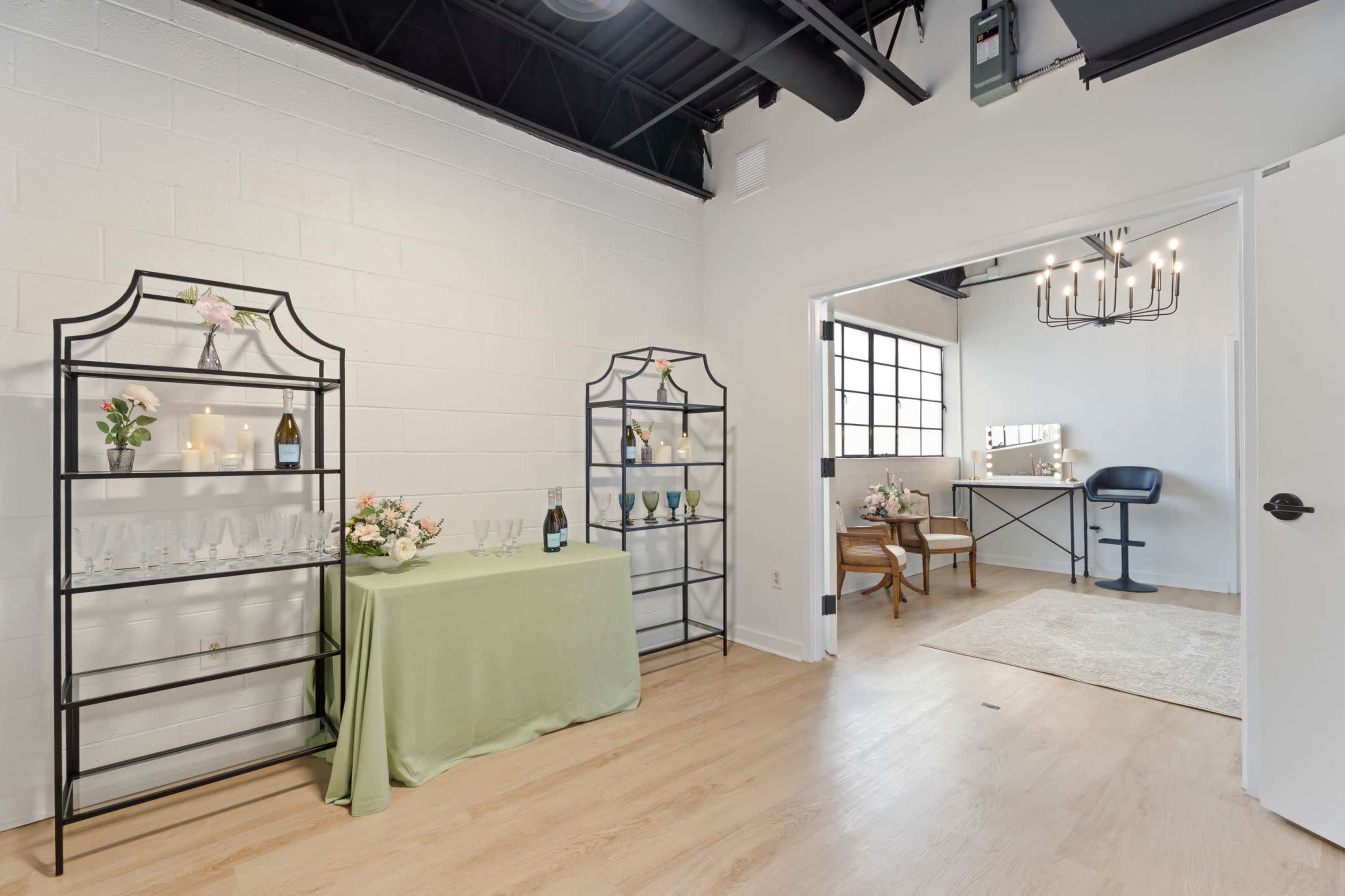 The image shows a bright room featuring a green tablecloth, two metal shelves displaying wine bottles and glasses, and a styled vanity area with a chair and mirror.