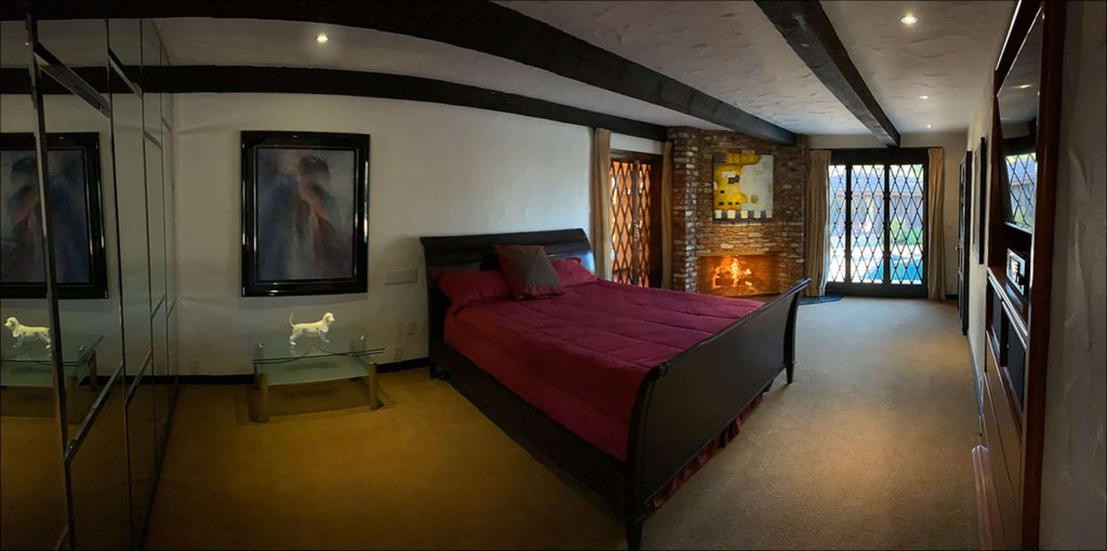 A cozy bedroom featuring a large bed with a maroon bedspread, artwork on the walls, and a stone fireplace.