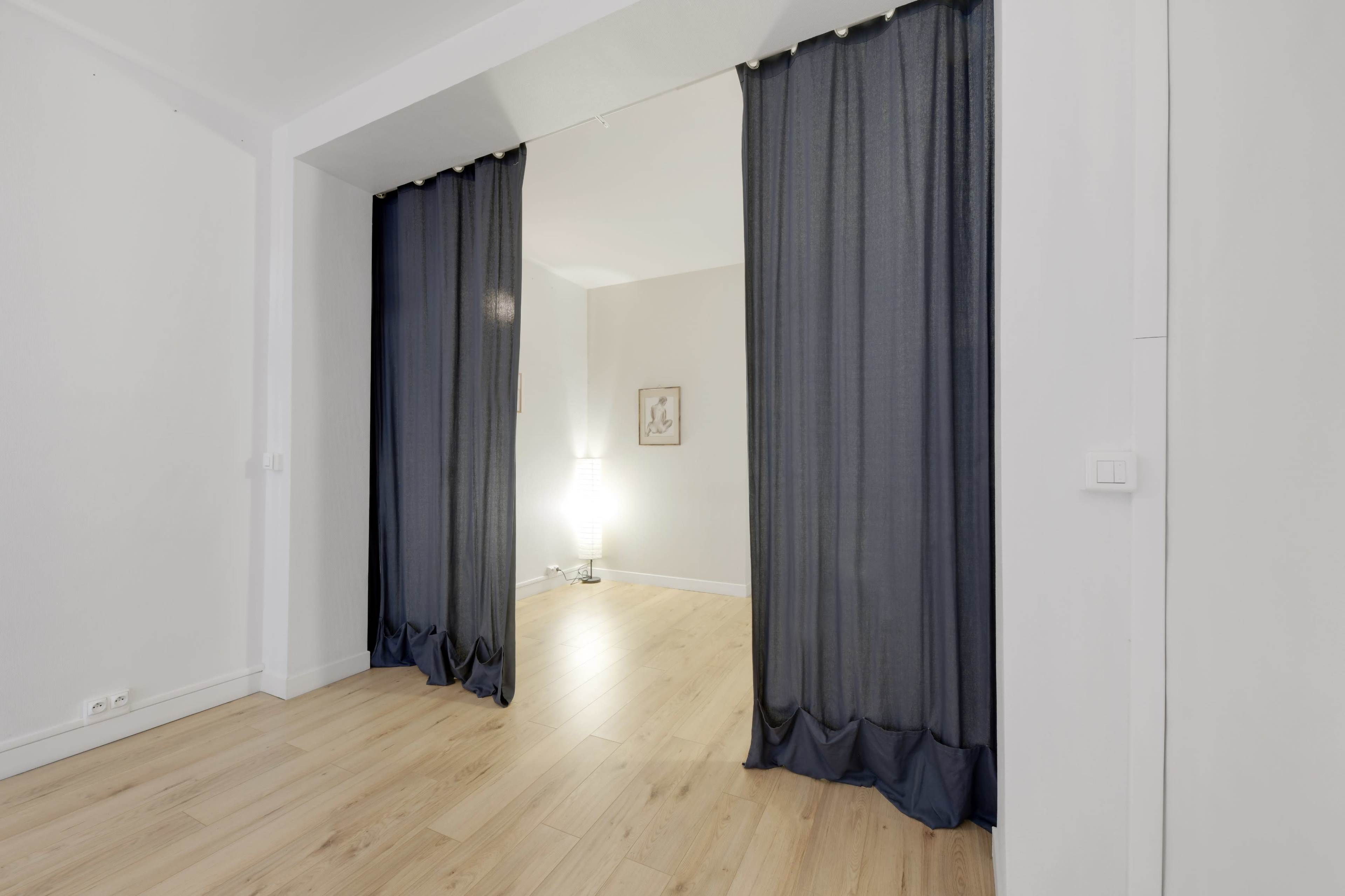 The image shows a partition made of dark curtains separating two areas in a room with wooden flooring and a lamp in the background.