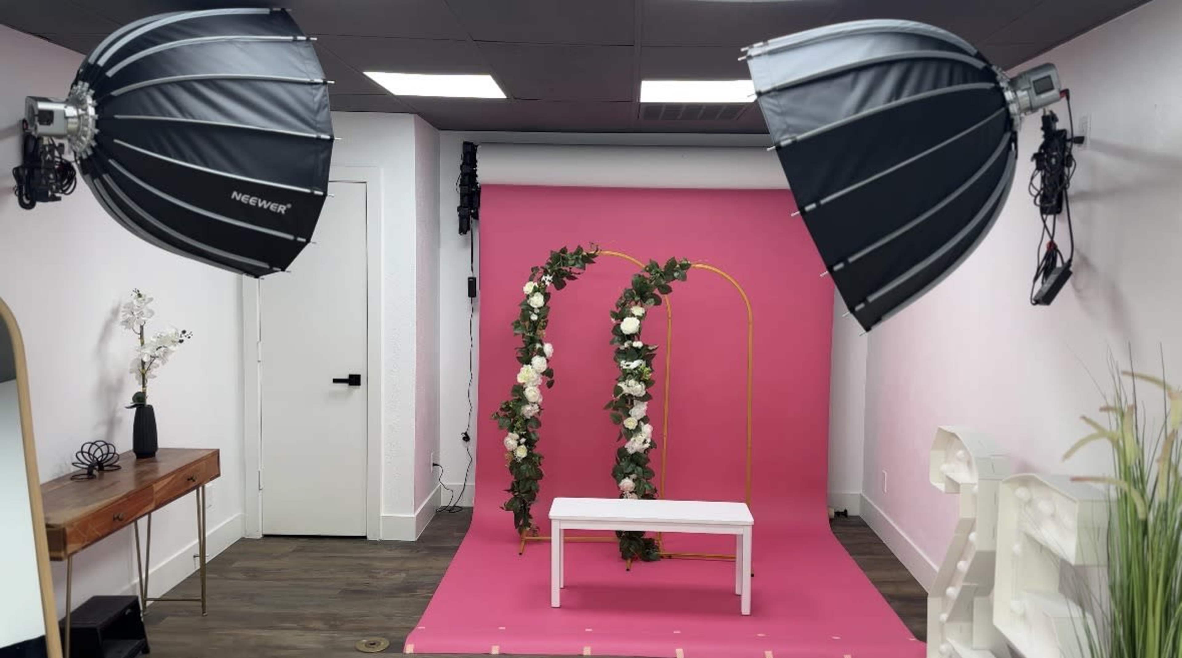 The image shows a brightly lit photography studio featuring a pink backdrop, a white bench, and decorative floral elements arranged on a golden arch.