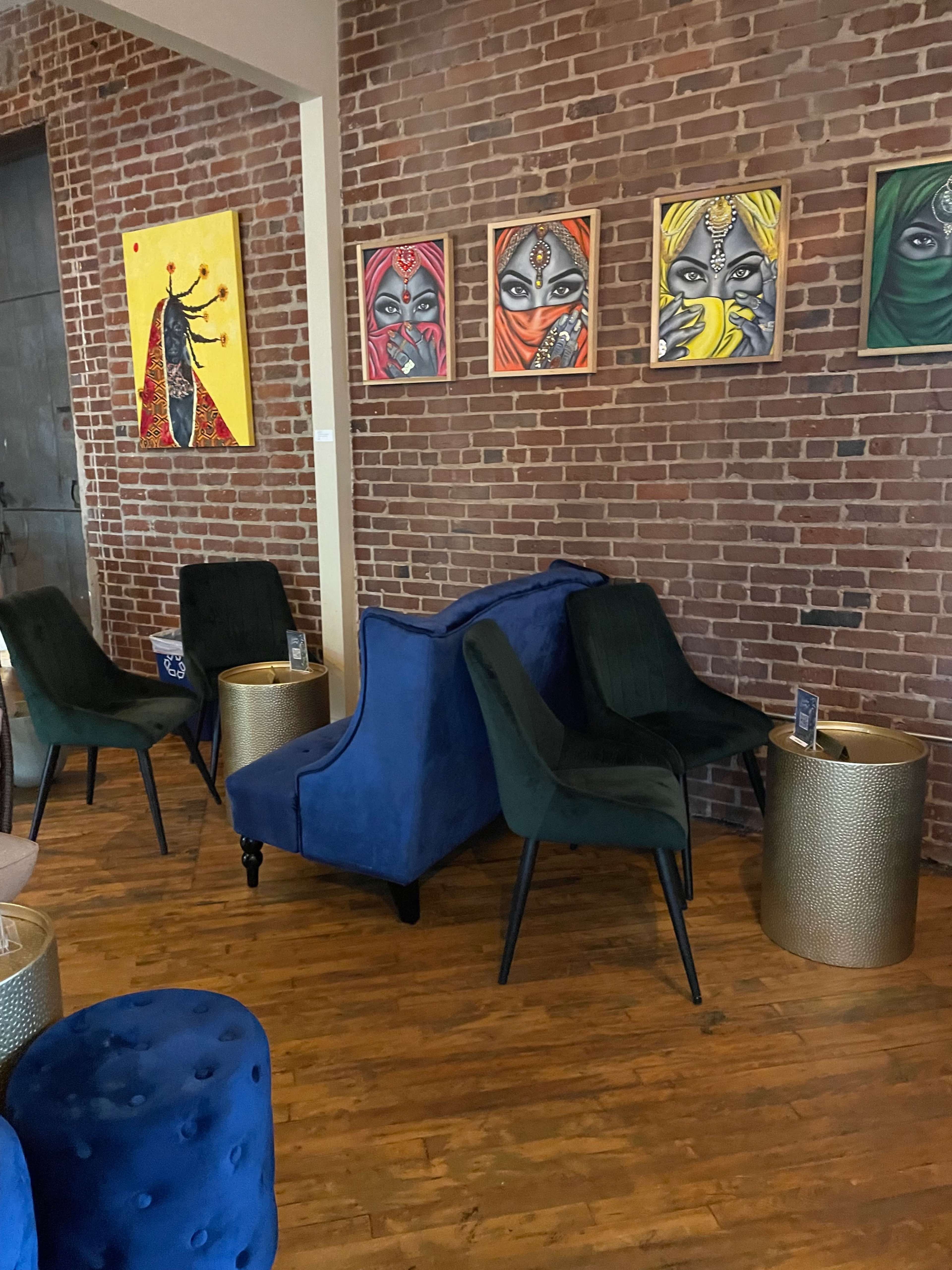 A seating area with blue and green chairs beside gold accent tables, against a backdrop of a brick wall adorned with colorful framed artwork.