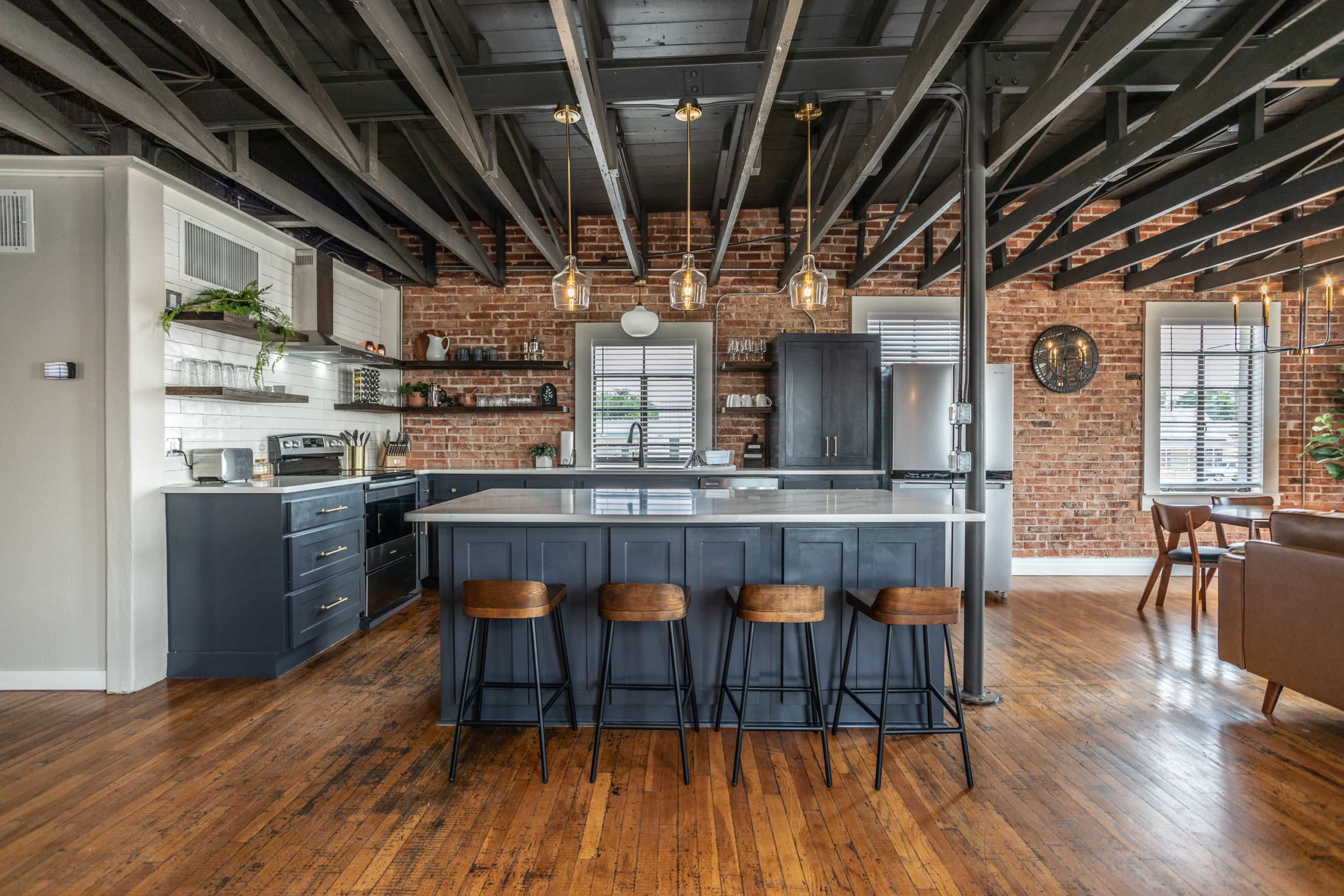 A modern kitchen with a central island, exposed wooden beams, brick walls, and a dining area in the background.