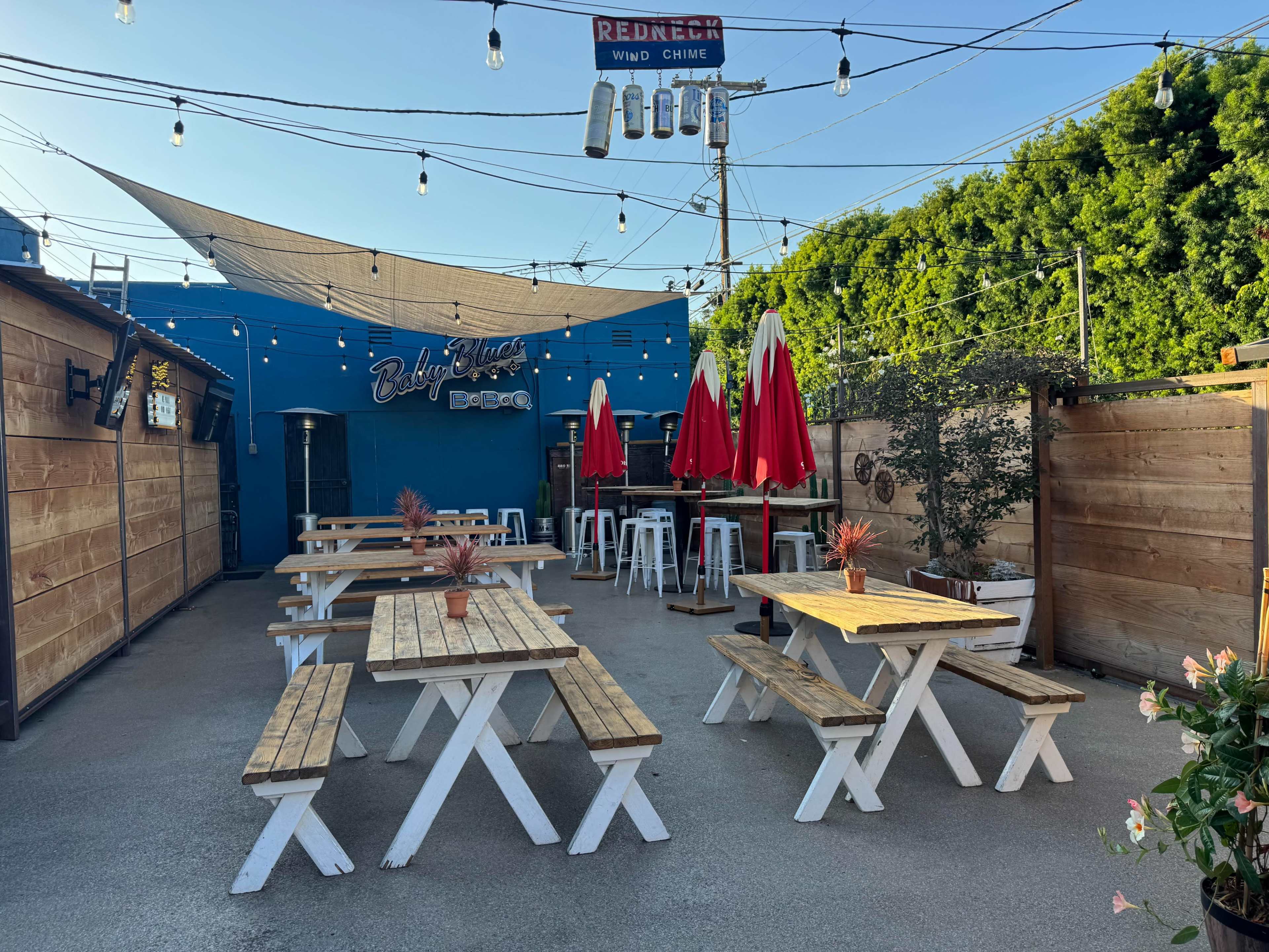 The outdoor seating area features wooden picnic tables with red and white umbrellas, surrounded by wooden fences and string lights against a blue wall.