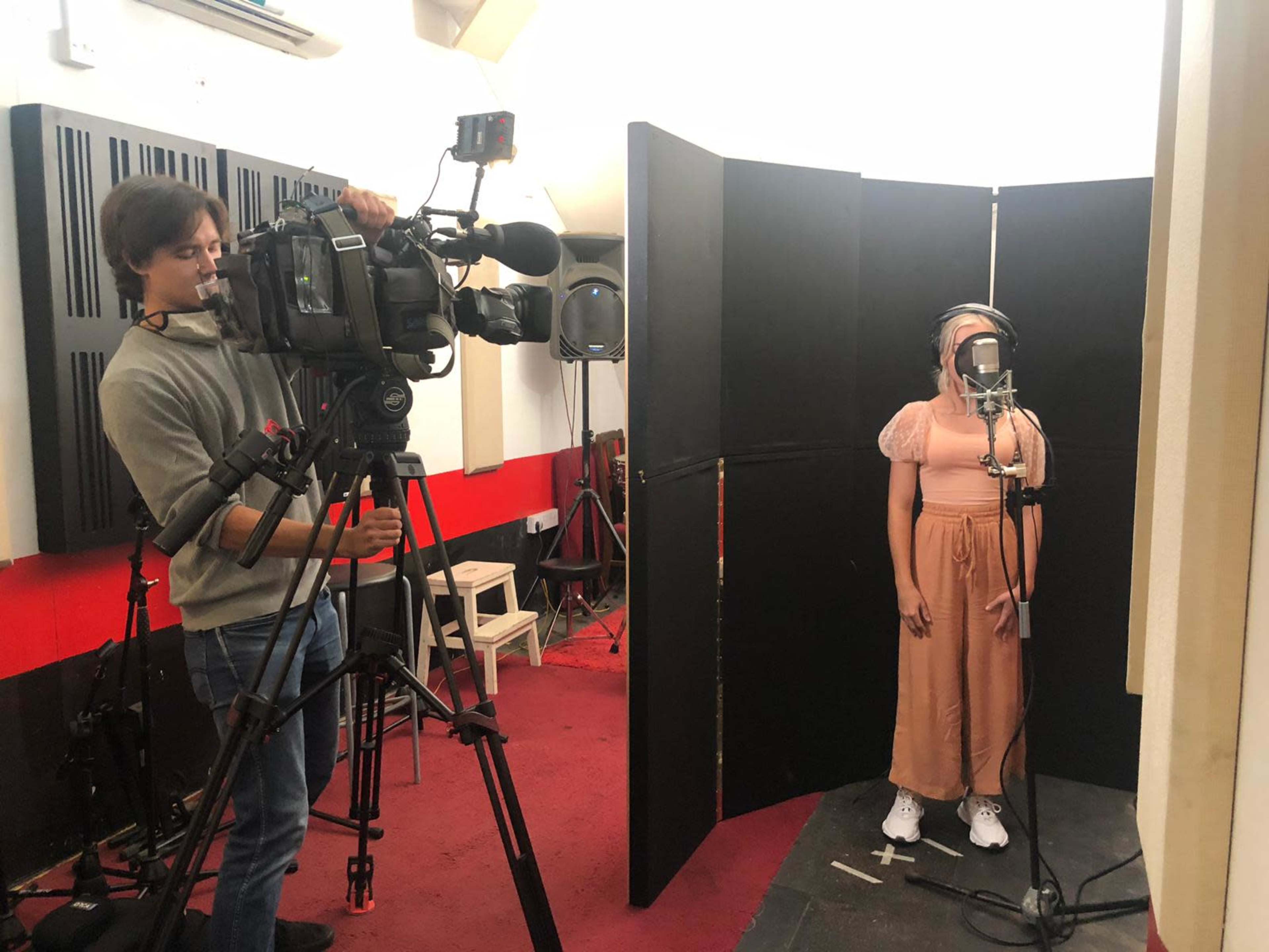 A person stands in front of a microphone in a recording studio while a cameraman films them.