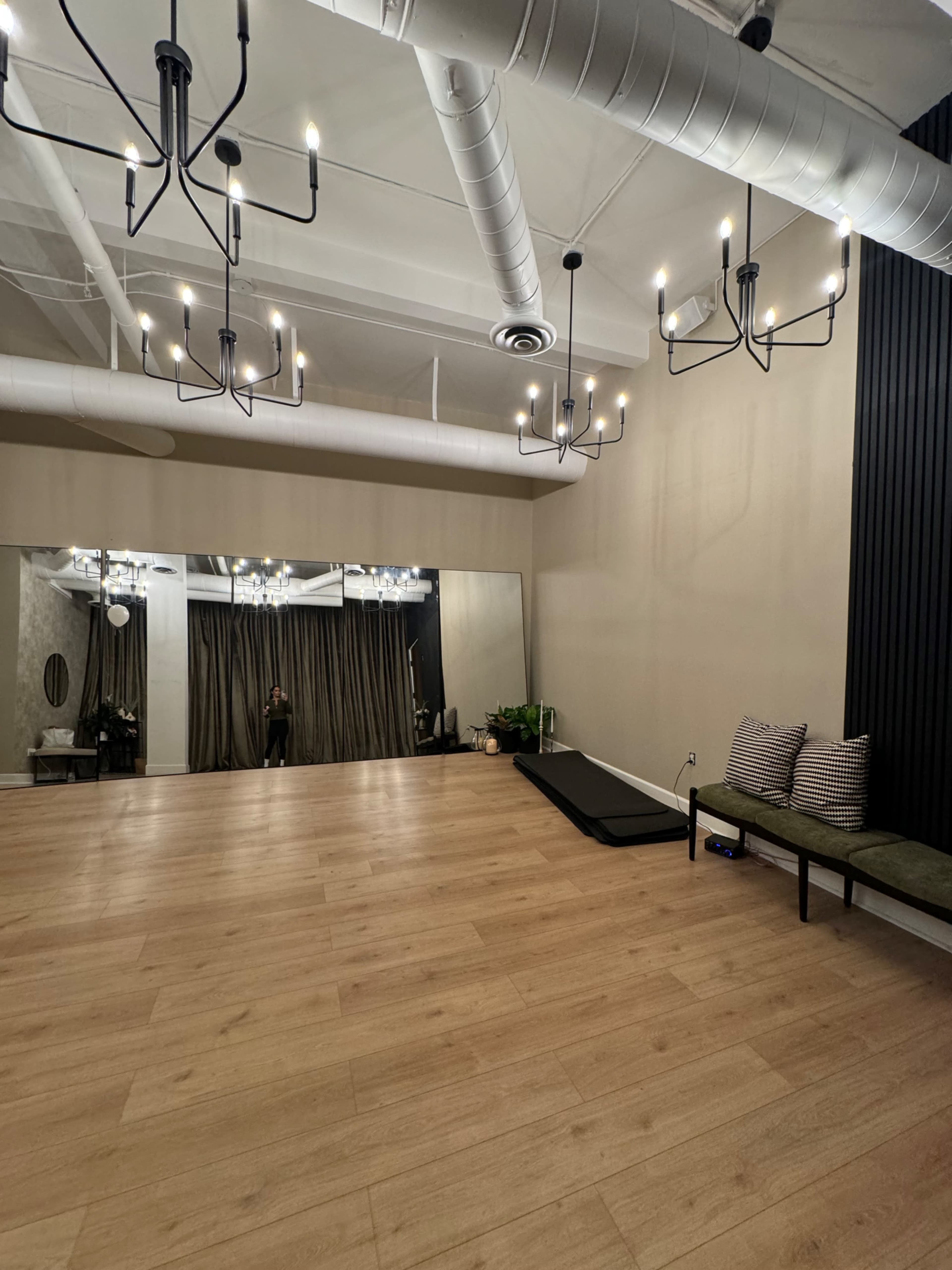 The image shows a spacious studio with wooden flooring, mirrored walls, and modern light fixtures hanging from the ceiling.