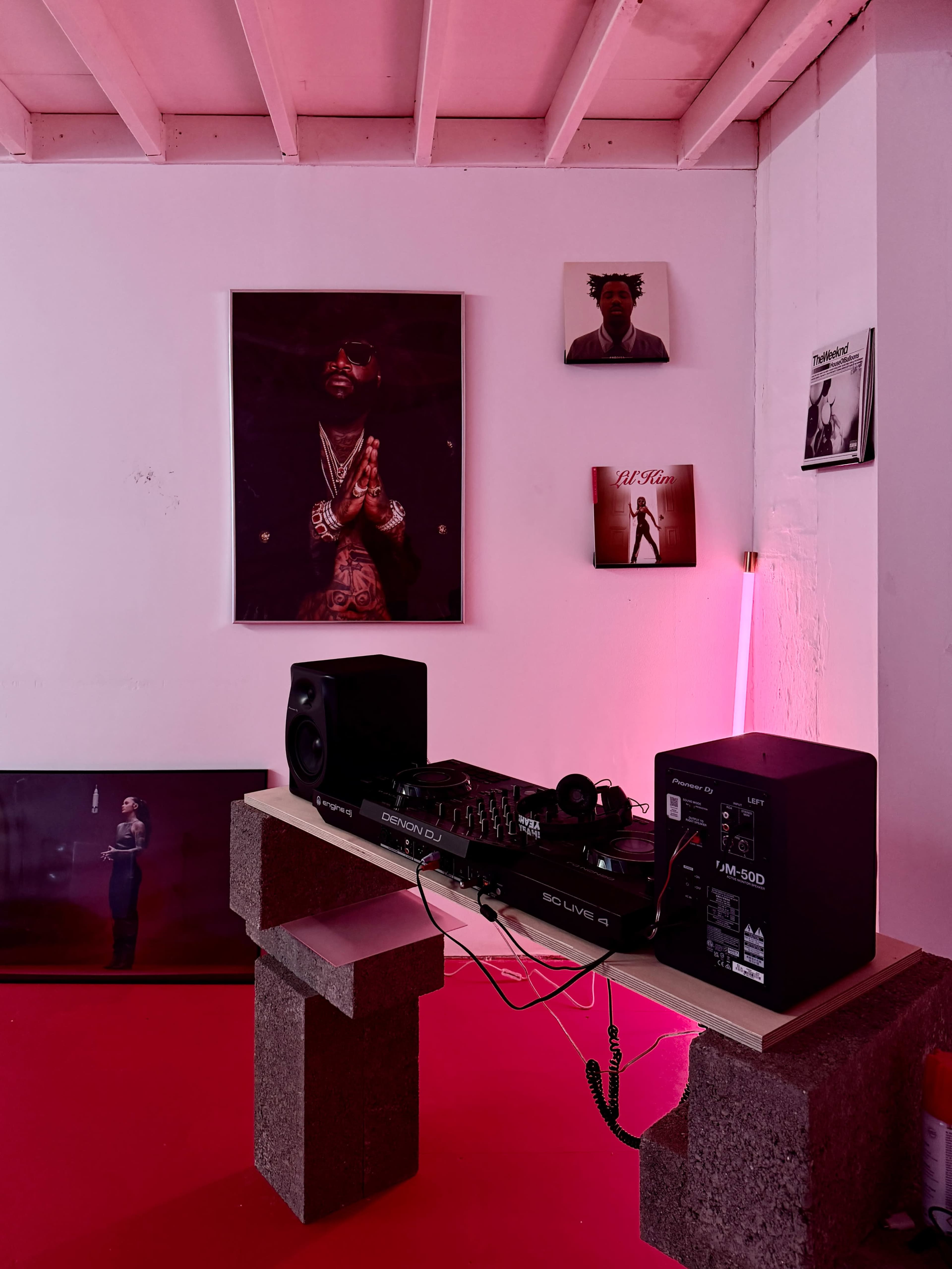 The image shows a music setup with a DJ controller and speakers on a table made of concrete blocks, surrounded by music-themed artwork on the walls and pink lighting.