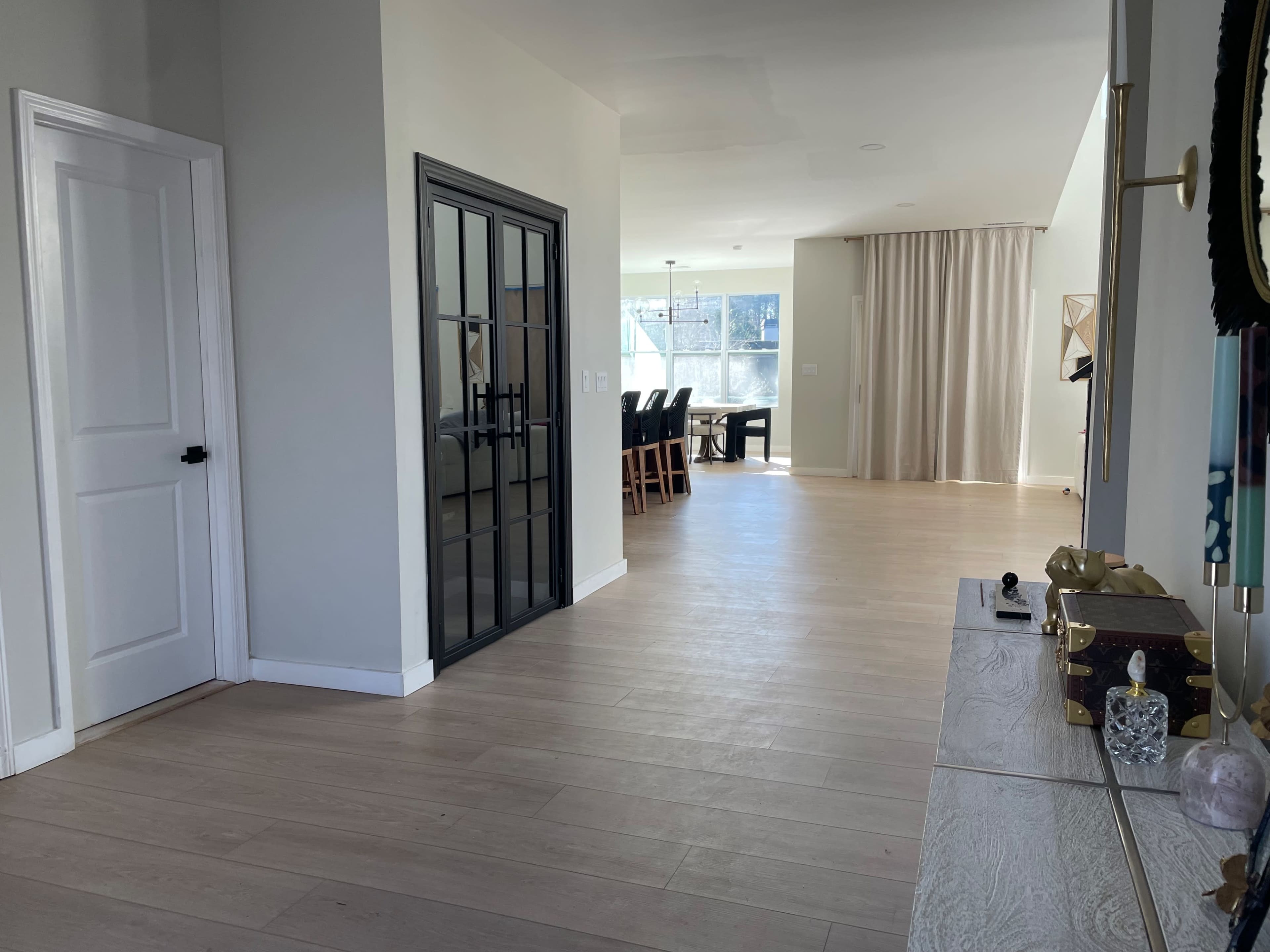 A hallway leading to an open living area with light-colored walls and a curtain-draped window.