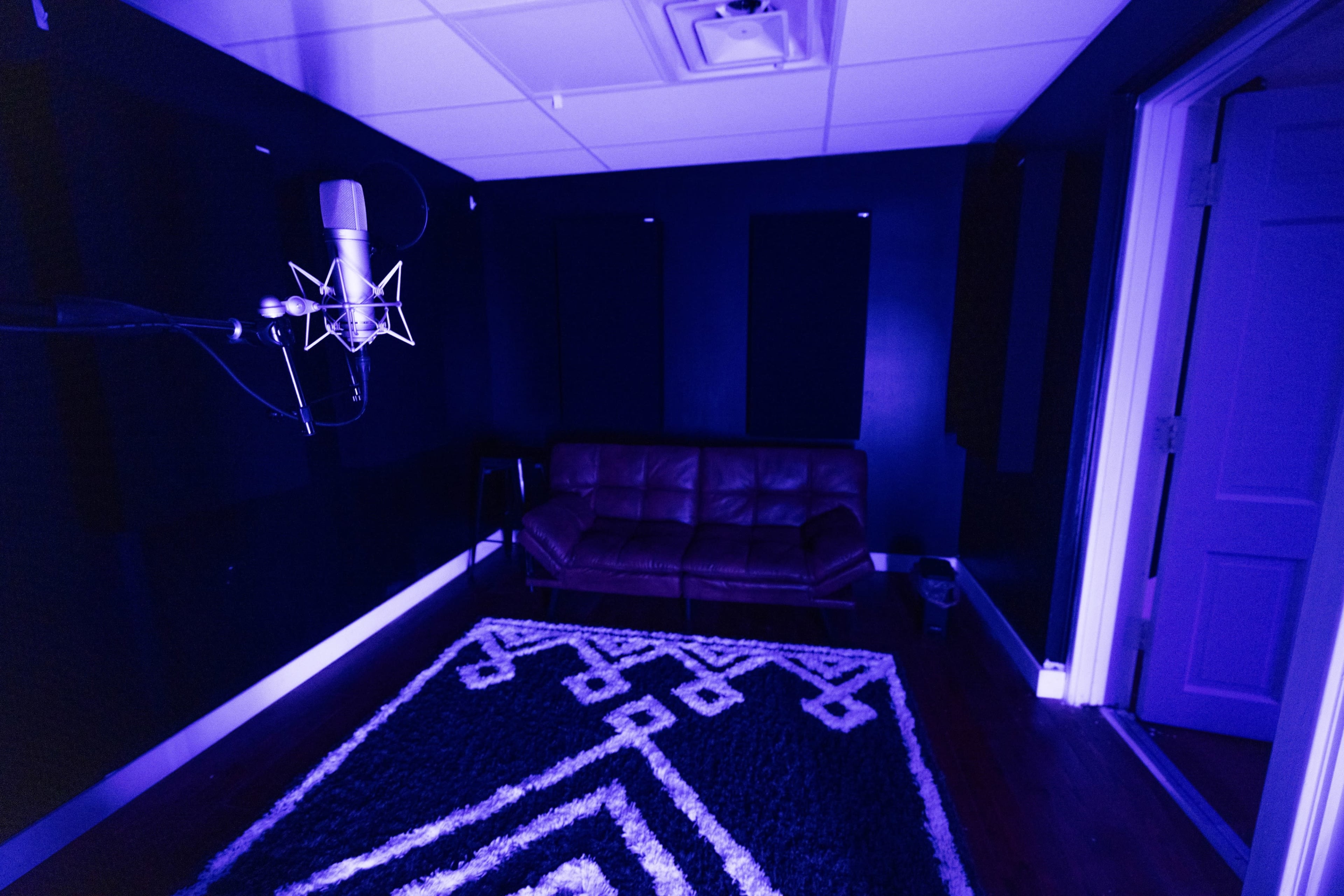 A recording studio features a microphone stand next to a dark leather couch and a patterned rug on the floor.