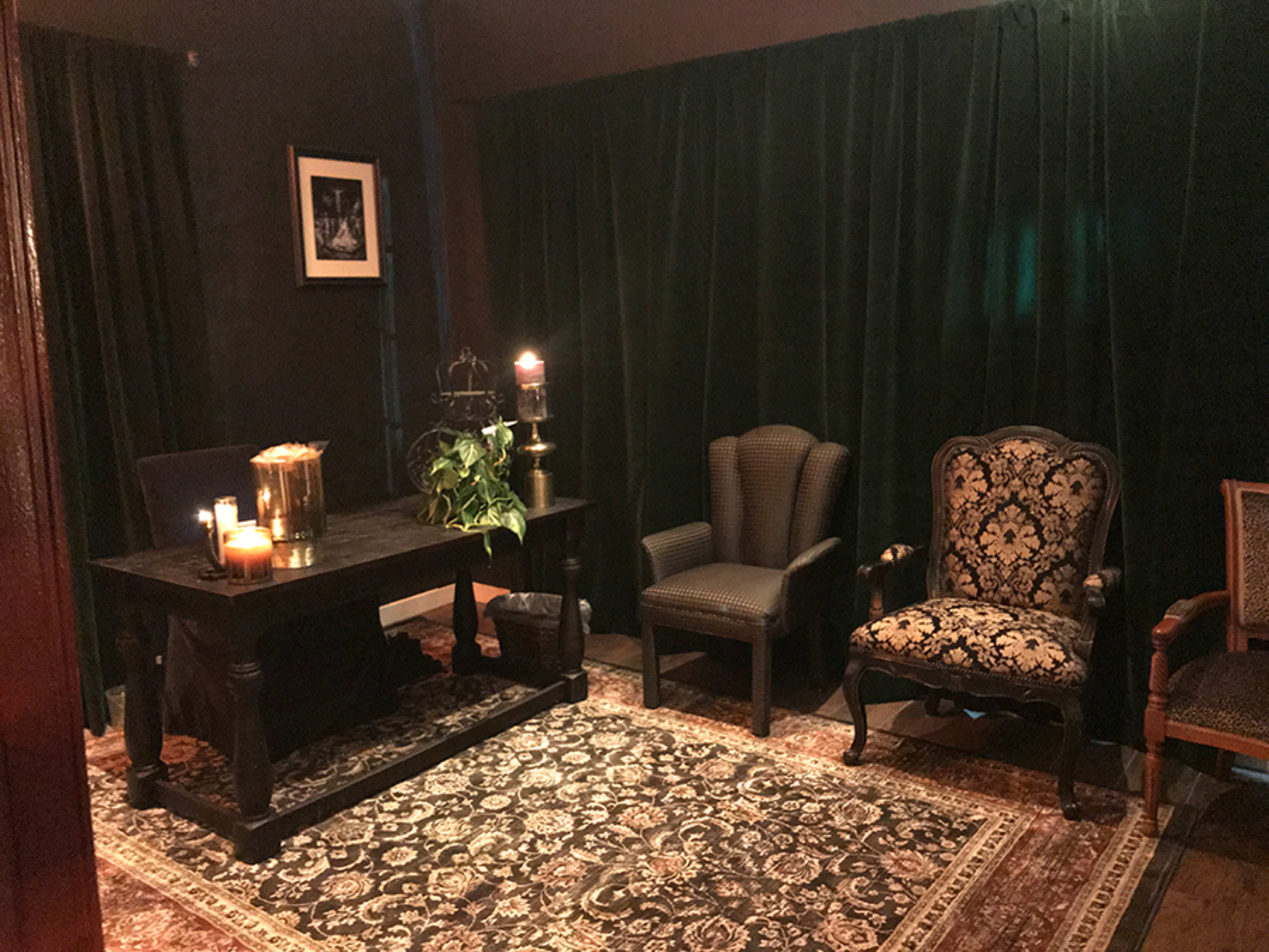 A dimly lit room with a dark green backdrop, featuring a table with candles, a potted plant, and two ornate chairs on a patterned rug.
