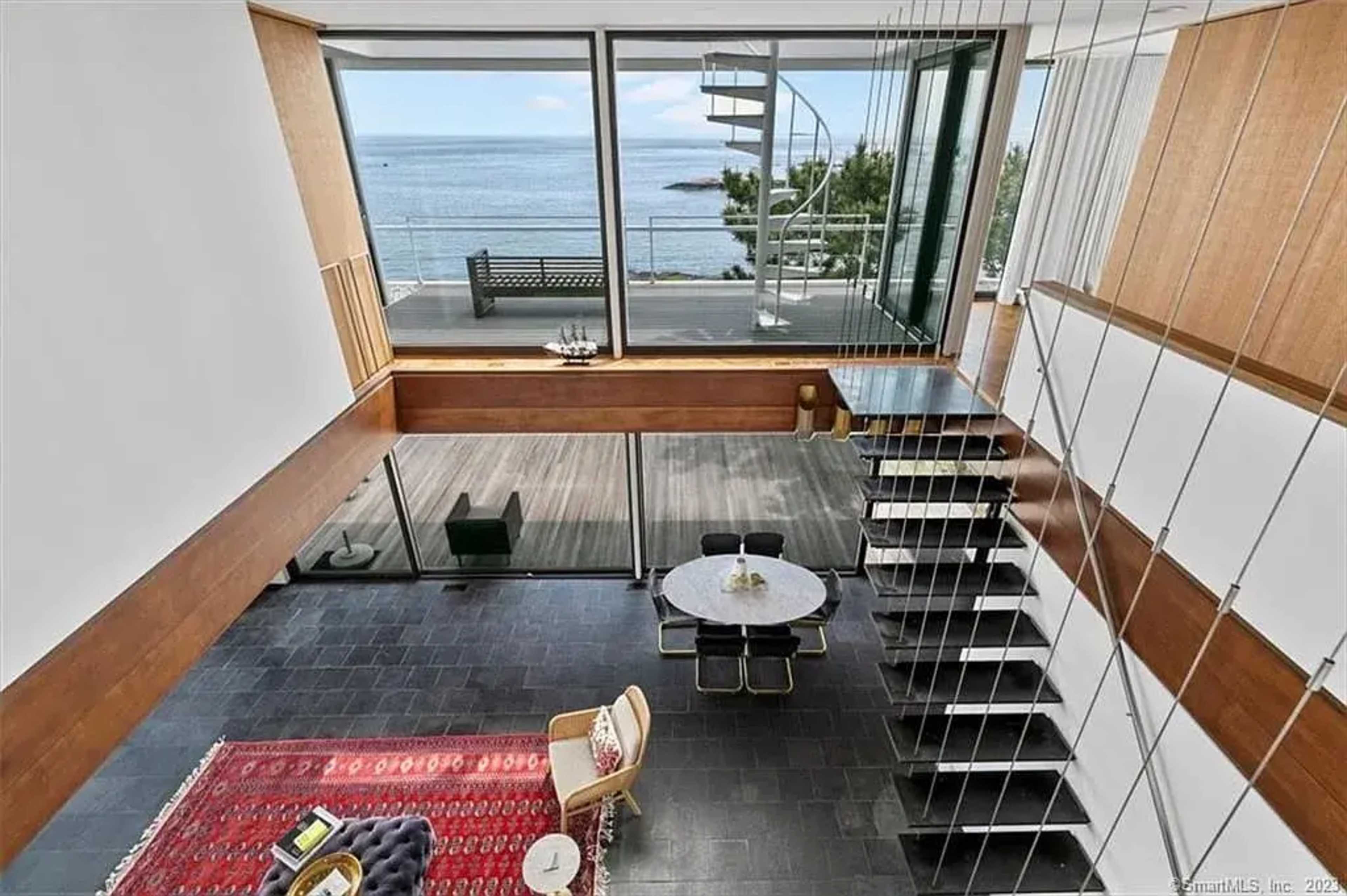 The image shows a modern interior space overlooking the ocean, featuring a staircase, a dining area with a round table, and large windows allowing natural light.