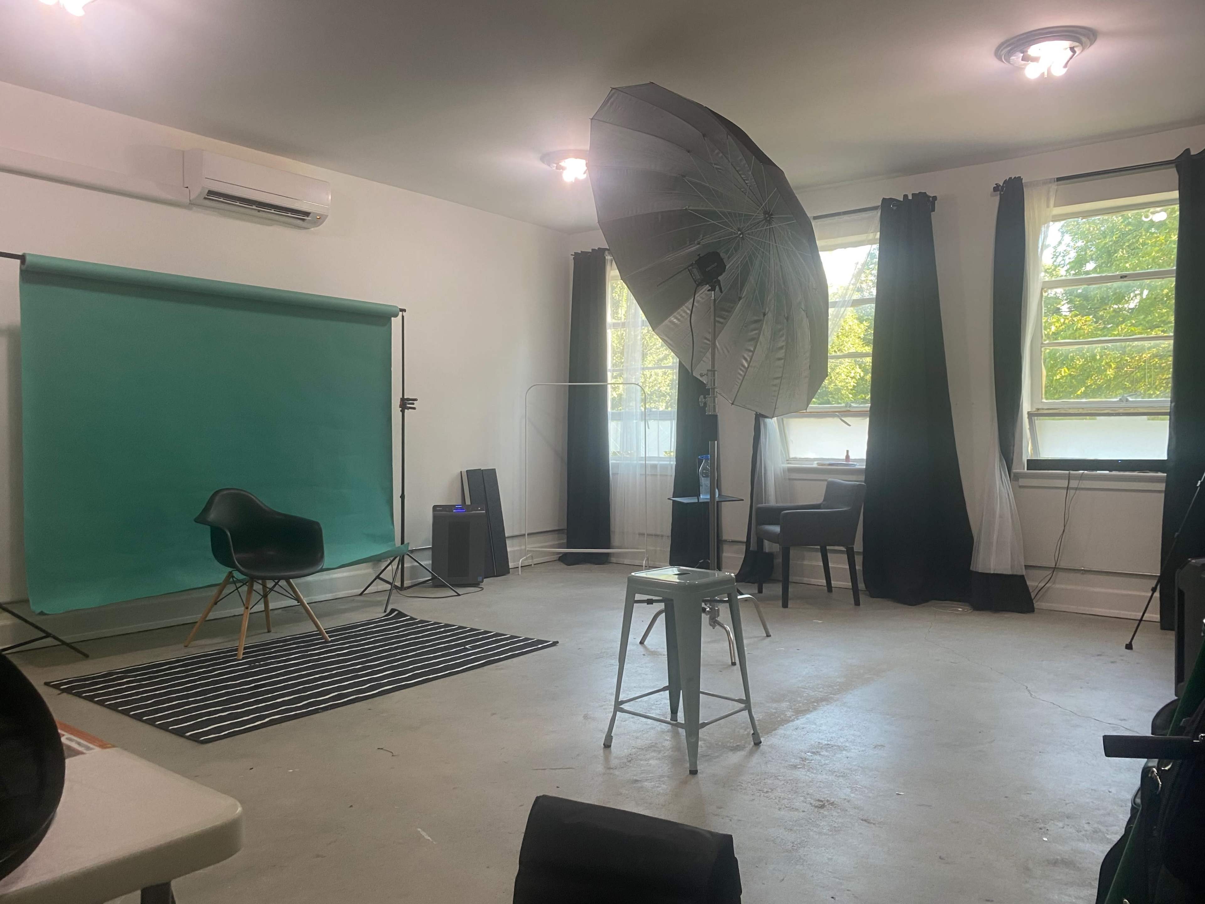 A photography studio featuring a green backdrop, a large softbox light, and minimalist furniture including a chair and a stool.