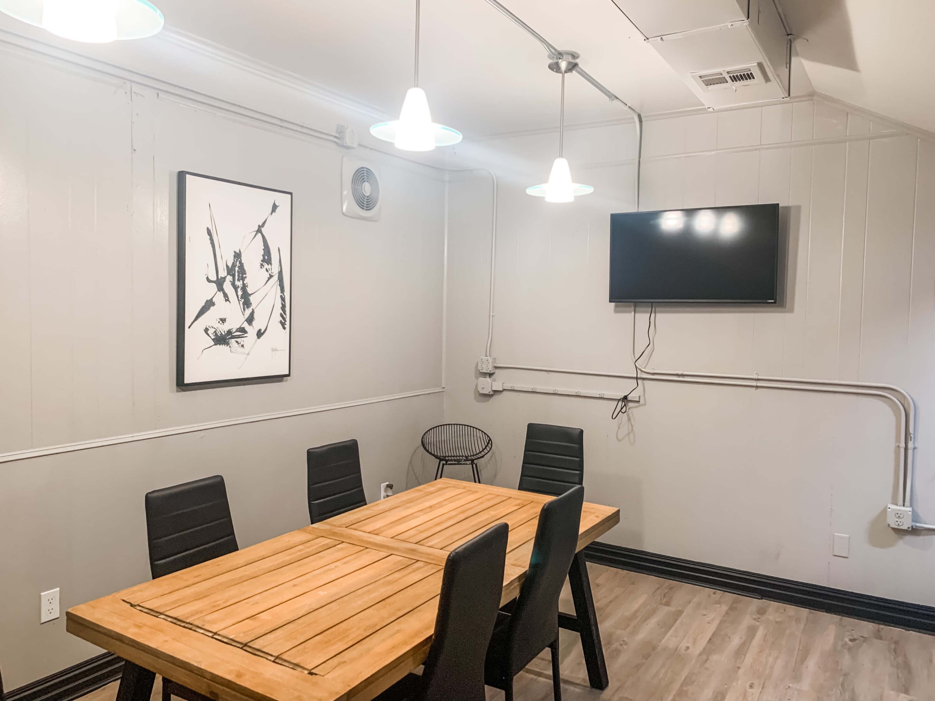 A modern conference room with a wooden table surrounded by six black chairs, a television mounted on the wall, and pendant lighting overhead.