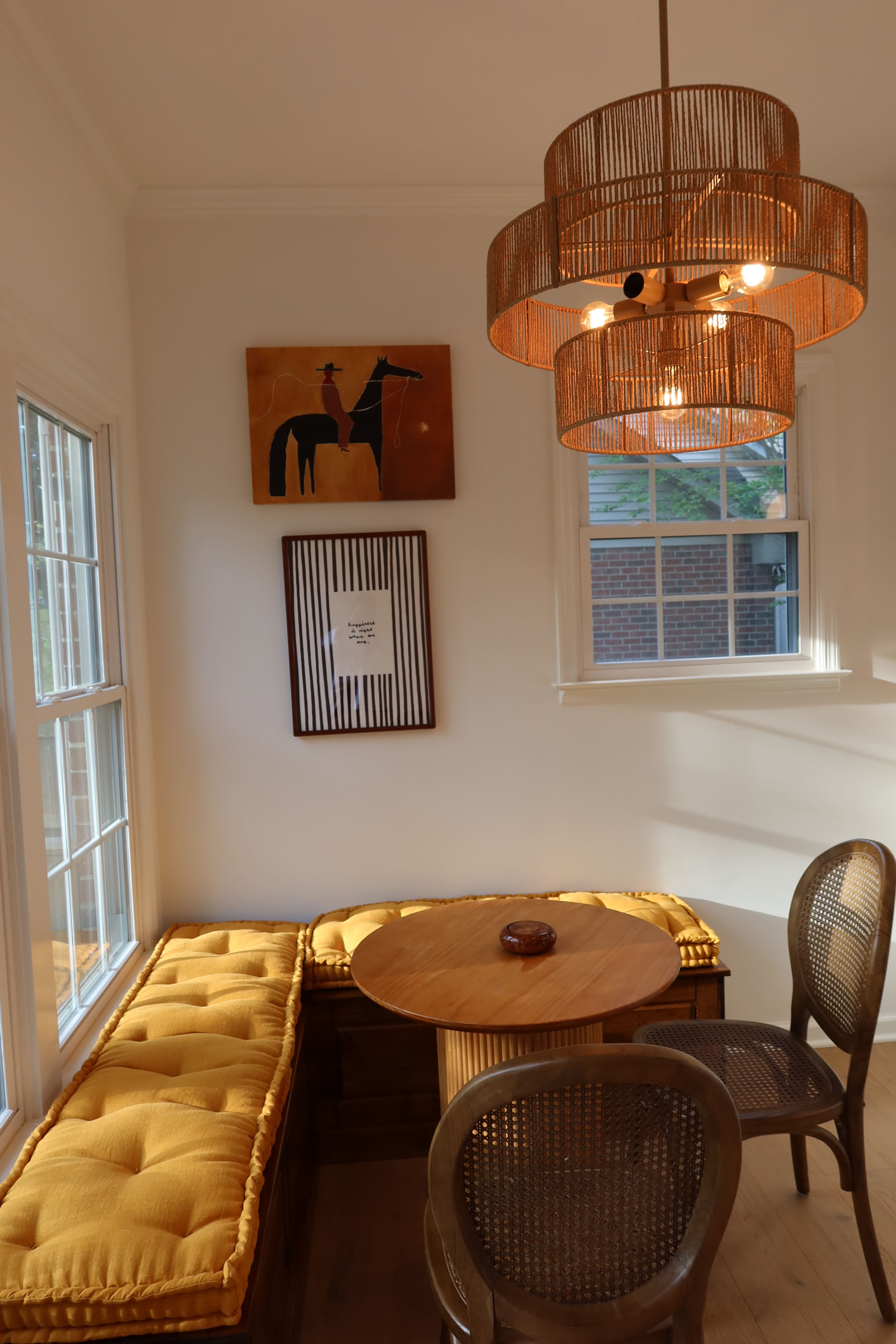The image shows a bright seating area with a round wooden table, two brown chairs, and a yellow cushioned bench beneath a lamp with a woven shade, accompanied by framed artwork on the walls.