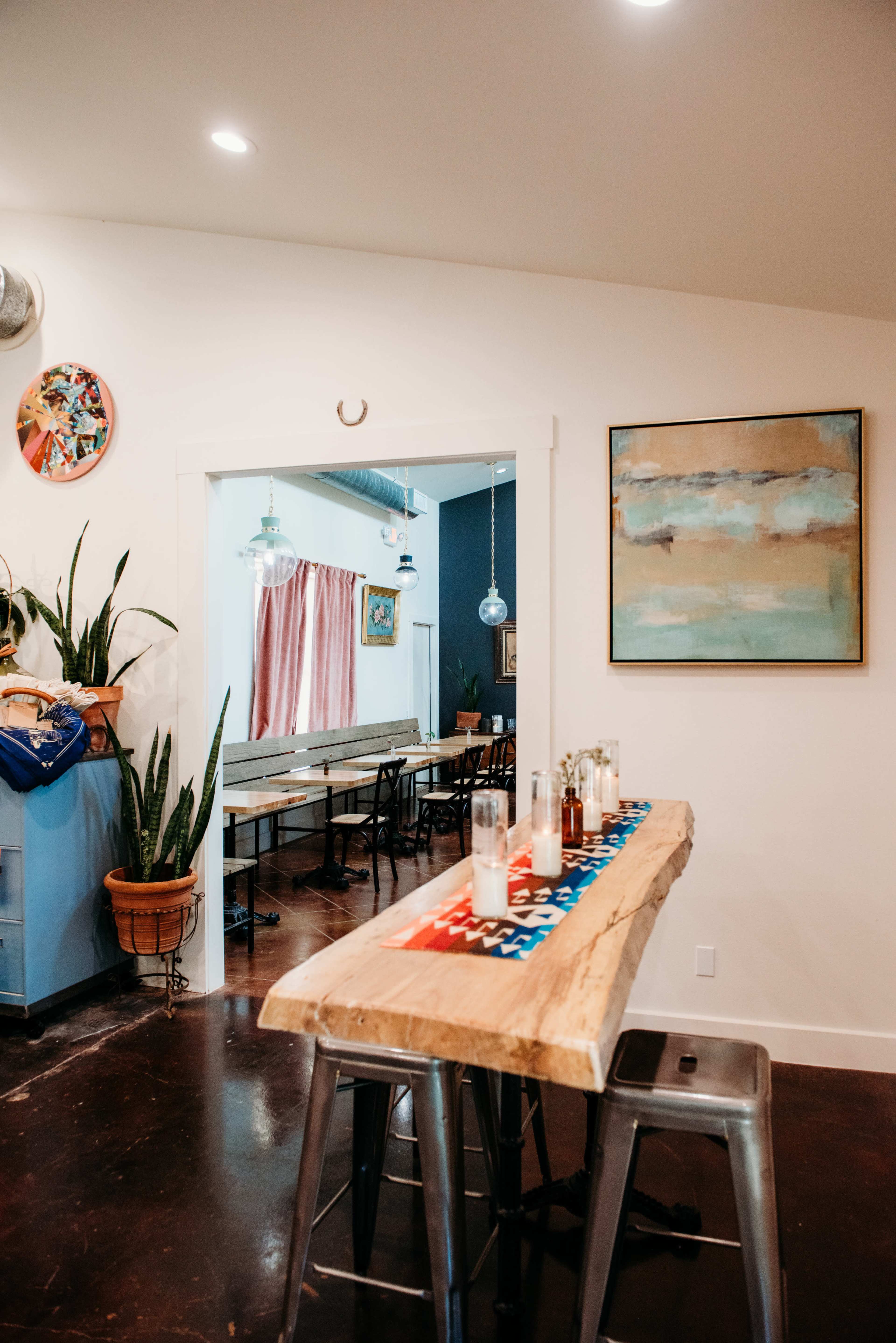 A dining area with a rustic wooden bar table, metal stools, and an open doorway leading to a room with tables and pink curtains.