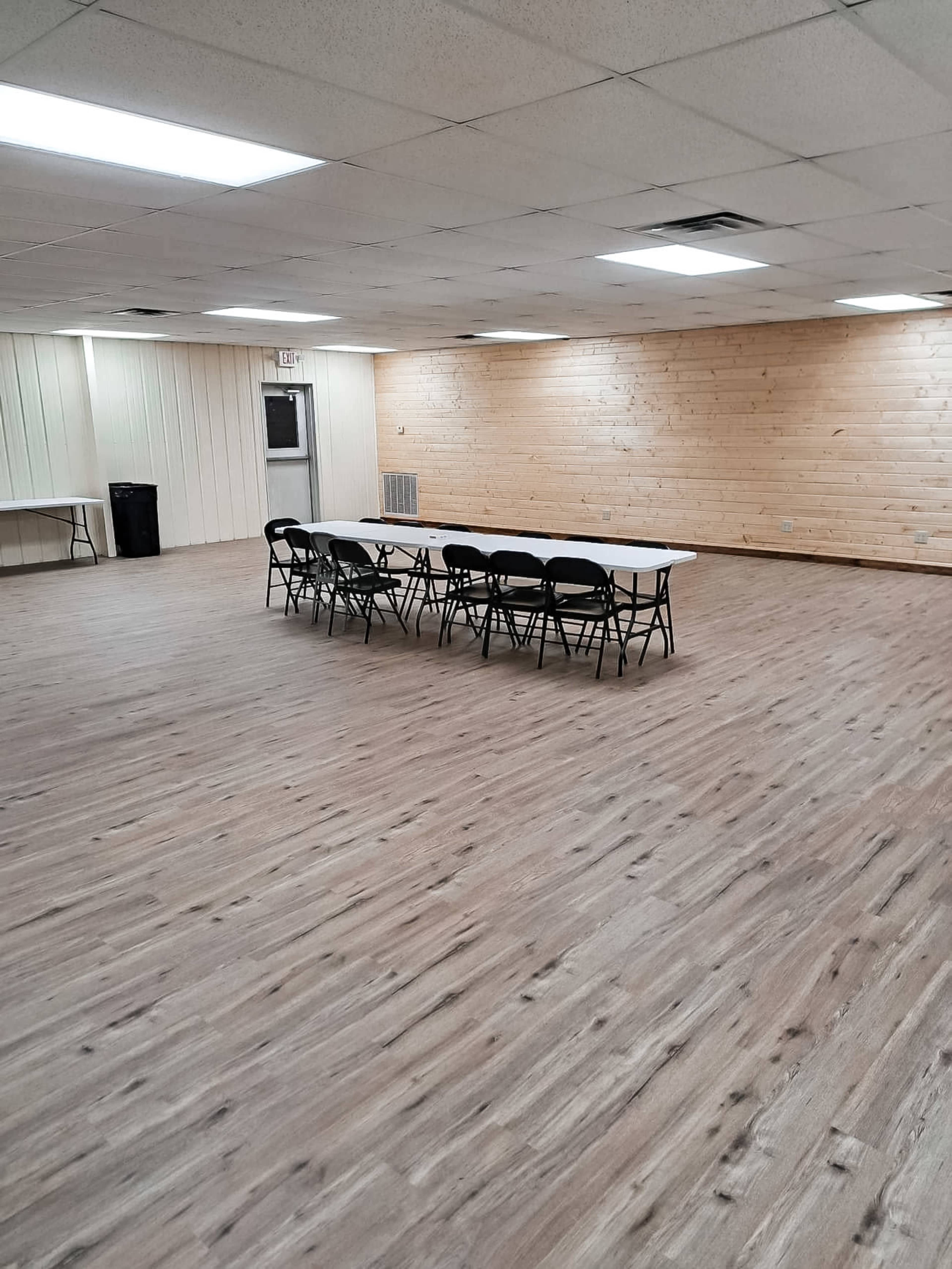 A large room features a long table surrounded by chairs, with wooden walls and a spacious, uncarpeted floor.