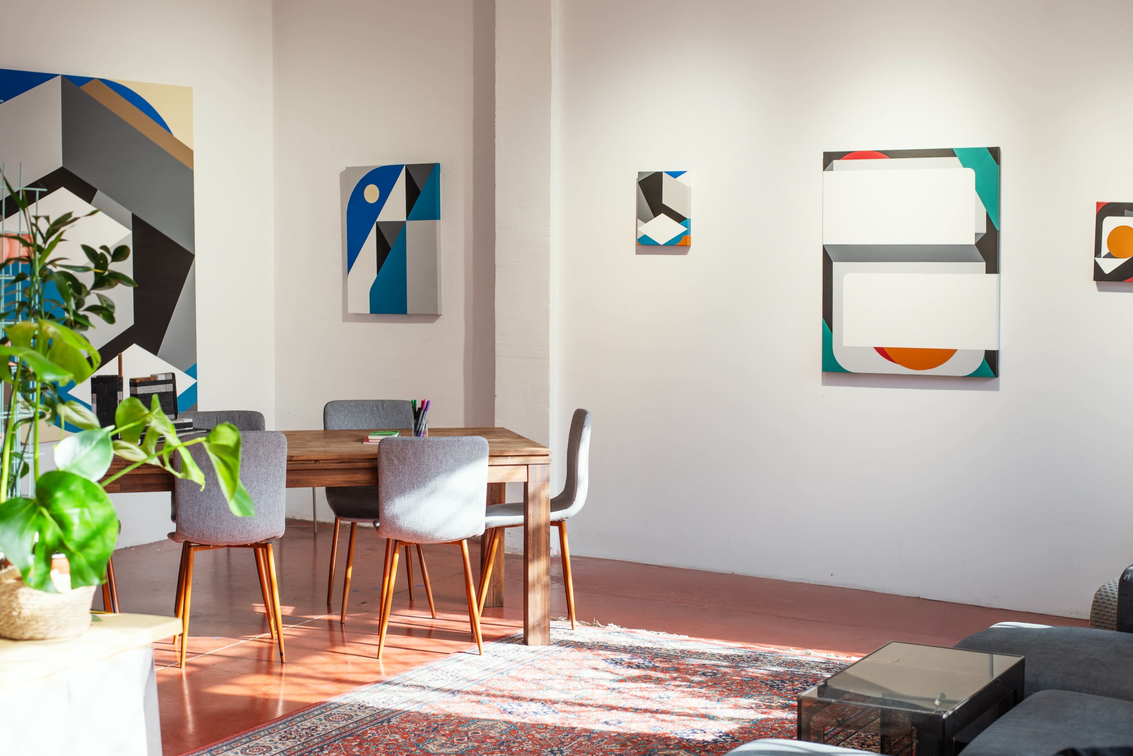 A bright, modern workspace features a wooden table surrounded by gray chairs, with colorful abstract artworks hanging on the walls.