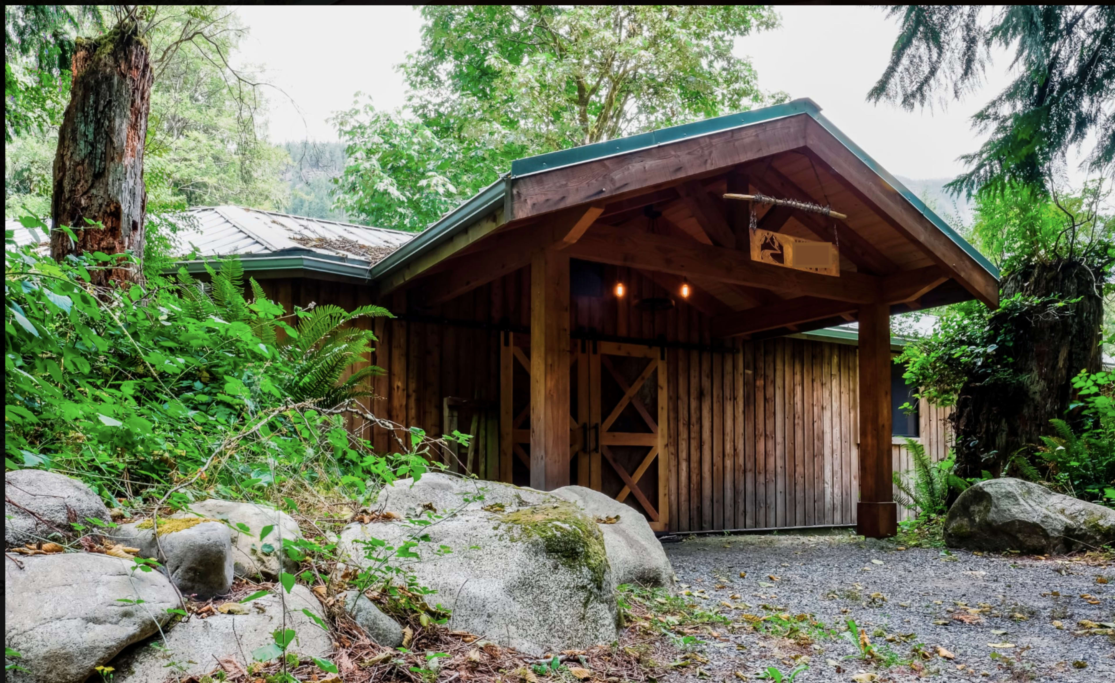 A wooden cabin with a slanted roof is nestled among greenery and boulders, featuring a covered entrance and rustic design elements.
