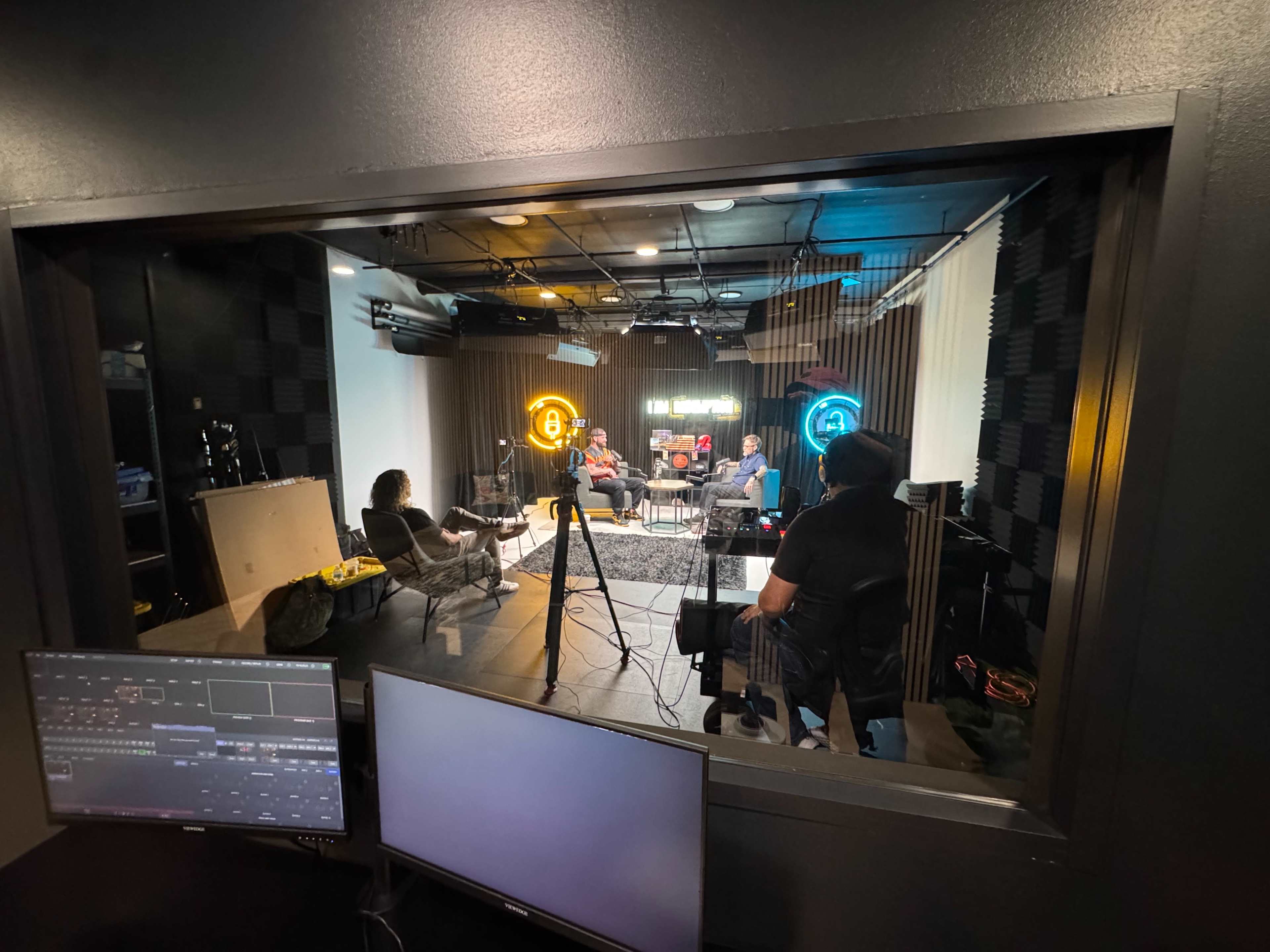 A group of people sits in a recording studio while being filmed, with colorful neon signs illuminating the background.