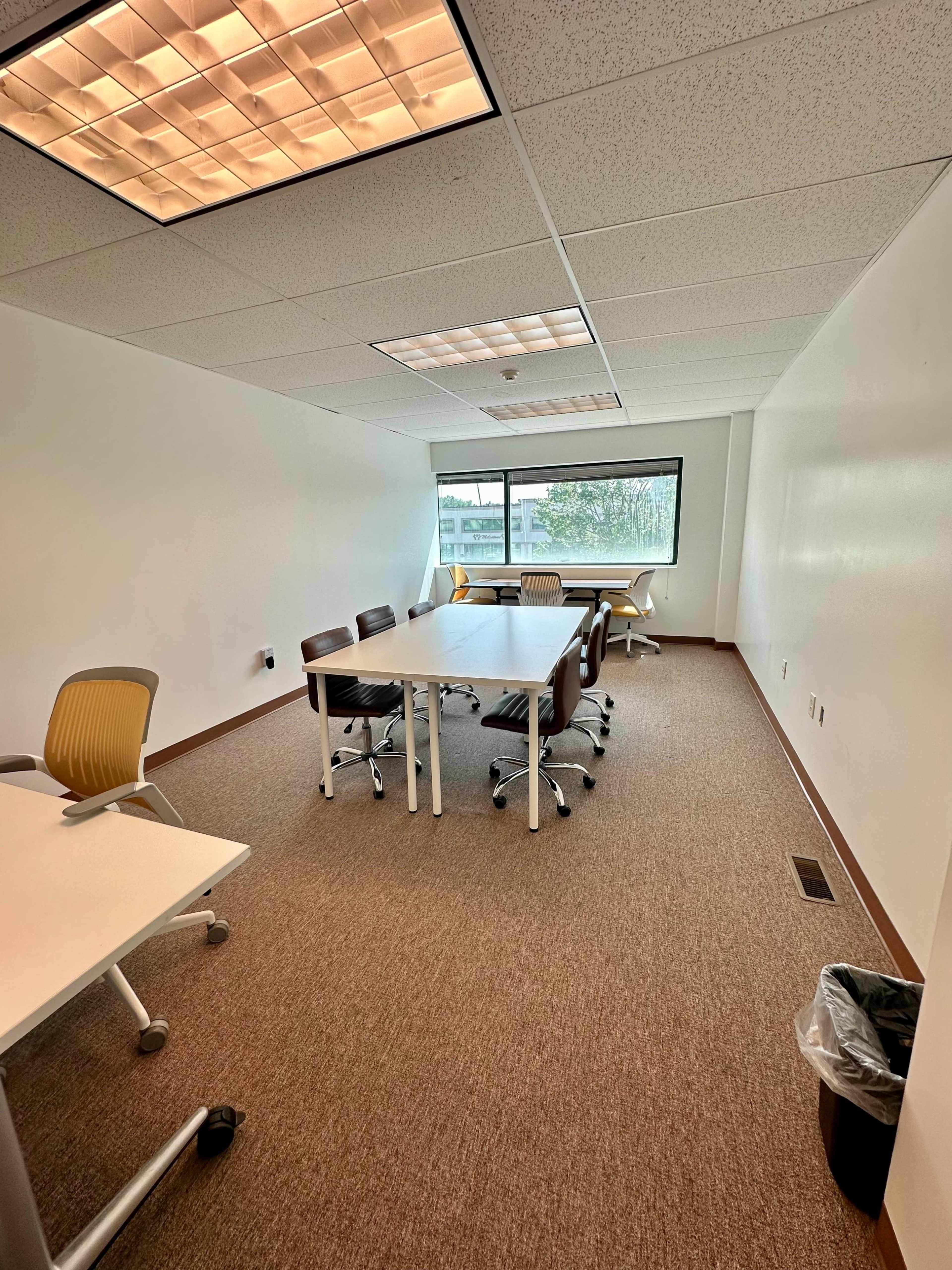 The image shows a modern office space with several tables and chairs arranged neatly, a large window, and a trash bin in the corner.