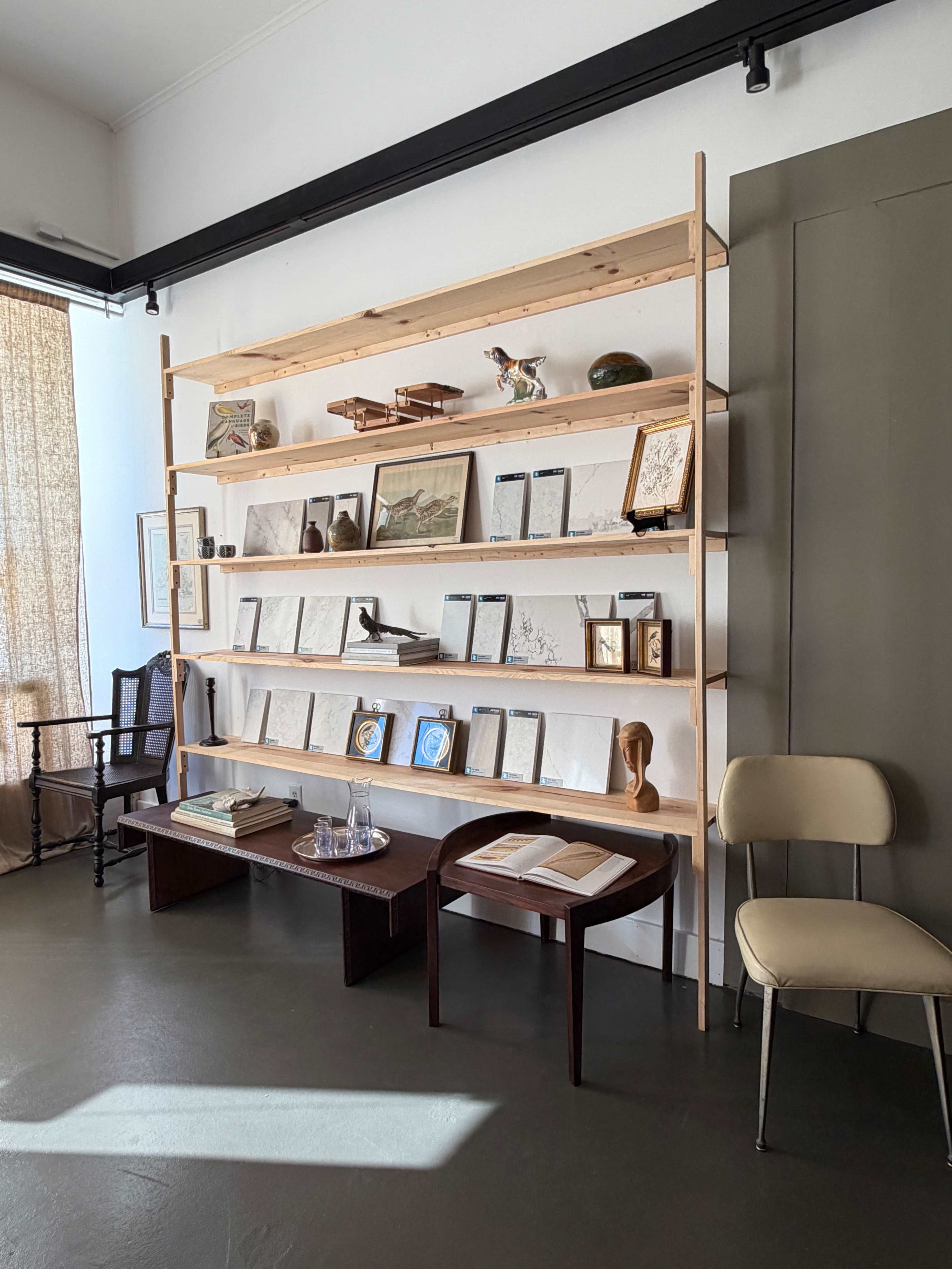 A wooden shelving unit holds a collection of framed artworks and books in a bright, minimalist room with a chair in the foreground.
