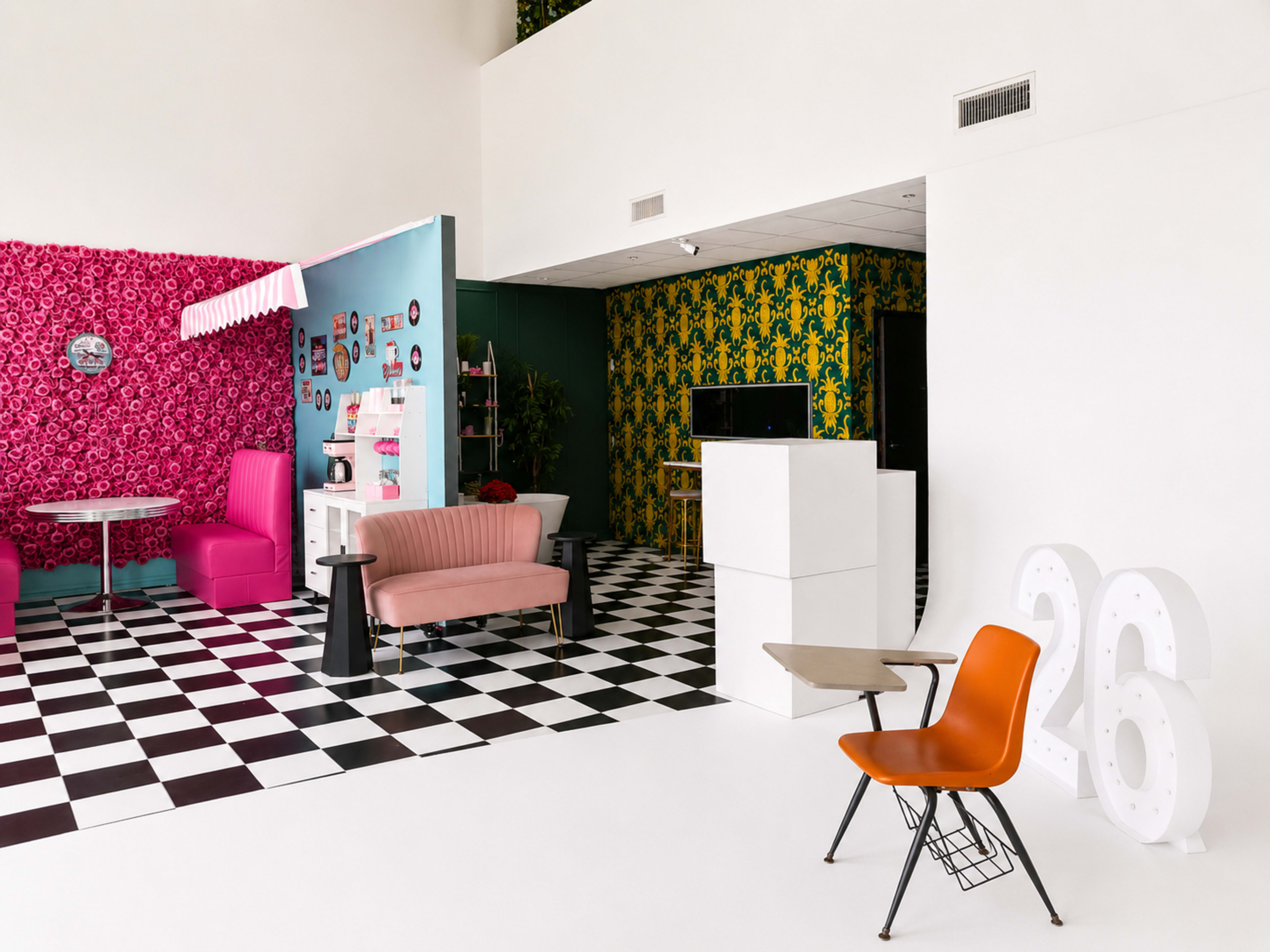 The image shows a brightly decorated room with contrasting wall patterns, featuring a pink seating area, a checkered floor, and various colorful decor elements.