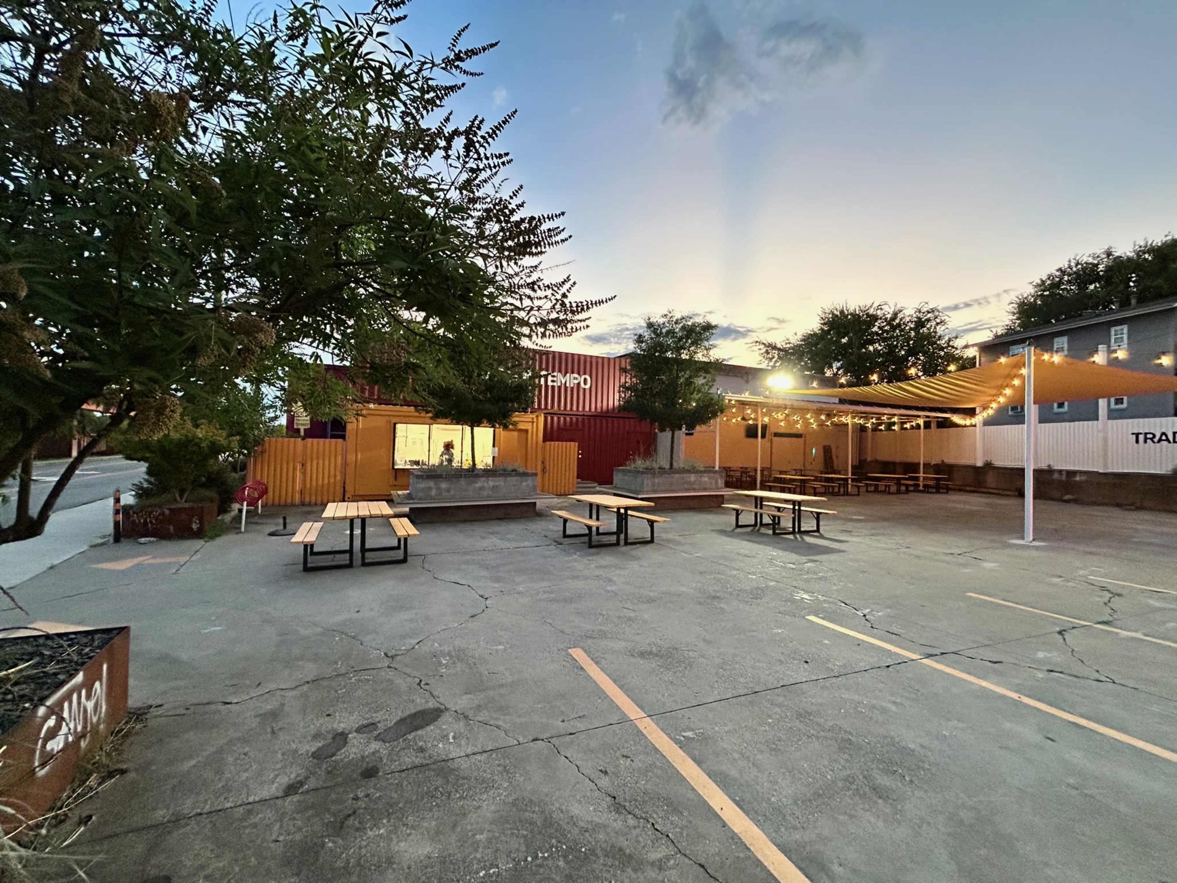 The image shows an outdoor dining area with several picnic tables set up in a gravel lot, illuminated by string lights amidst a backdrop of shipping container structures and trees at dusk.