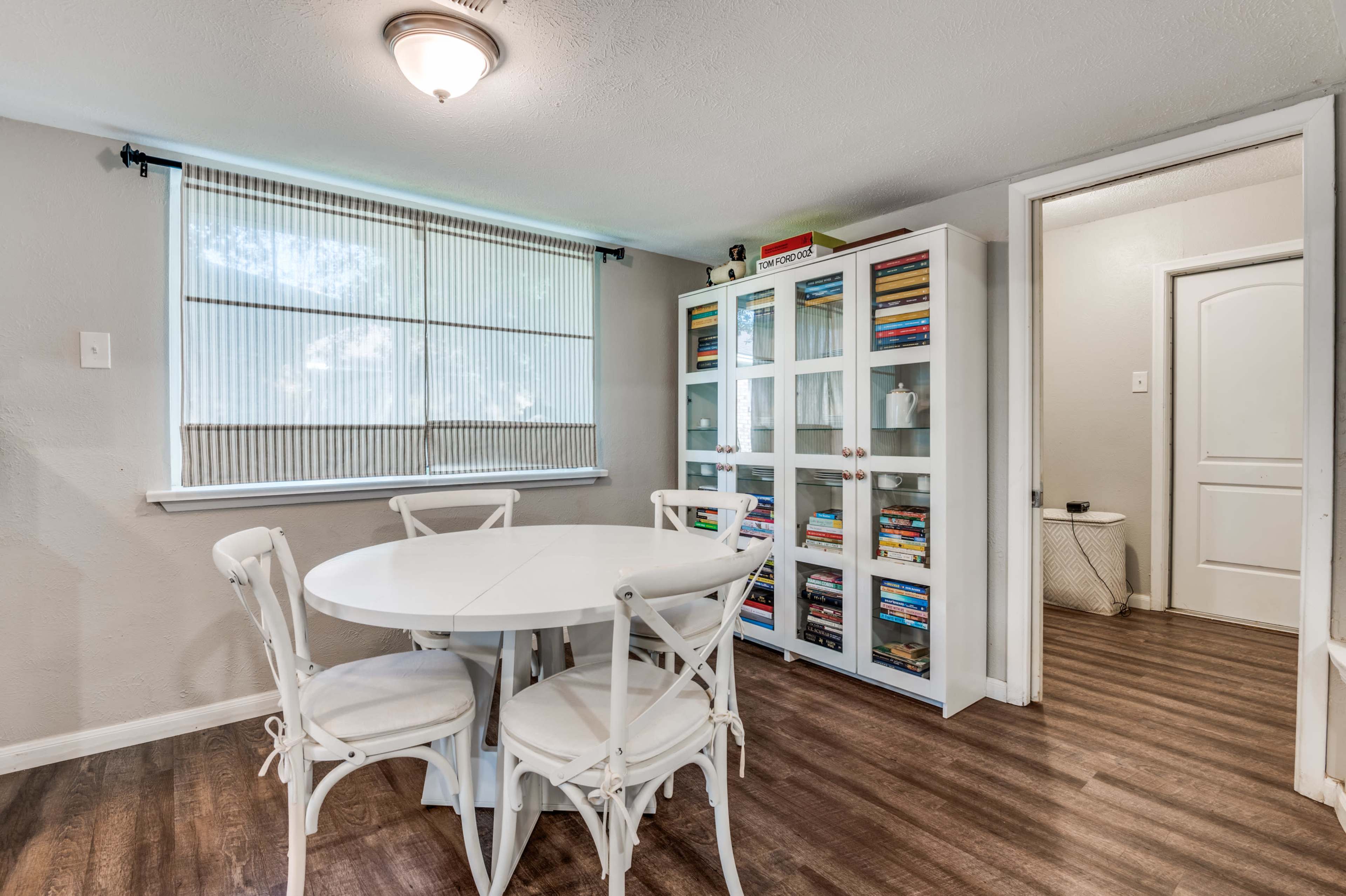 The image shows a small dining area with a round table surrounded by white chairs, a glass-front cabinet filled with books, and a door leading to another room.