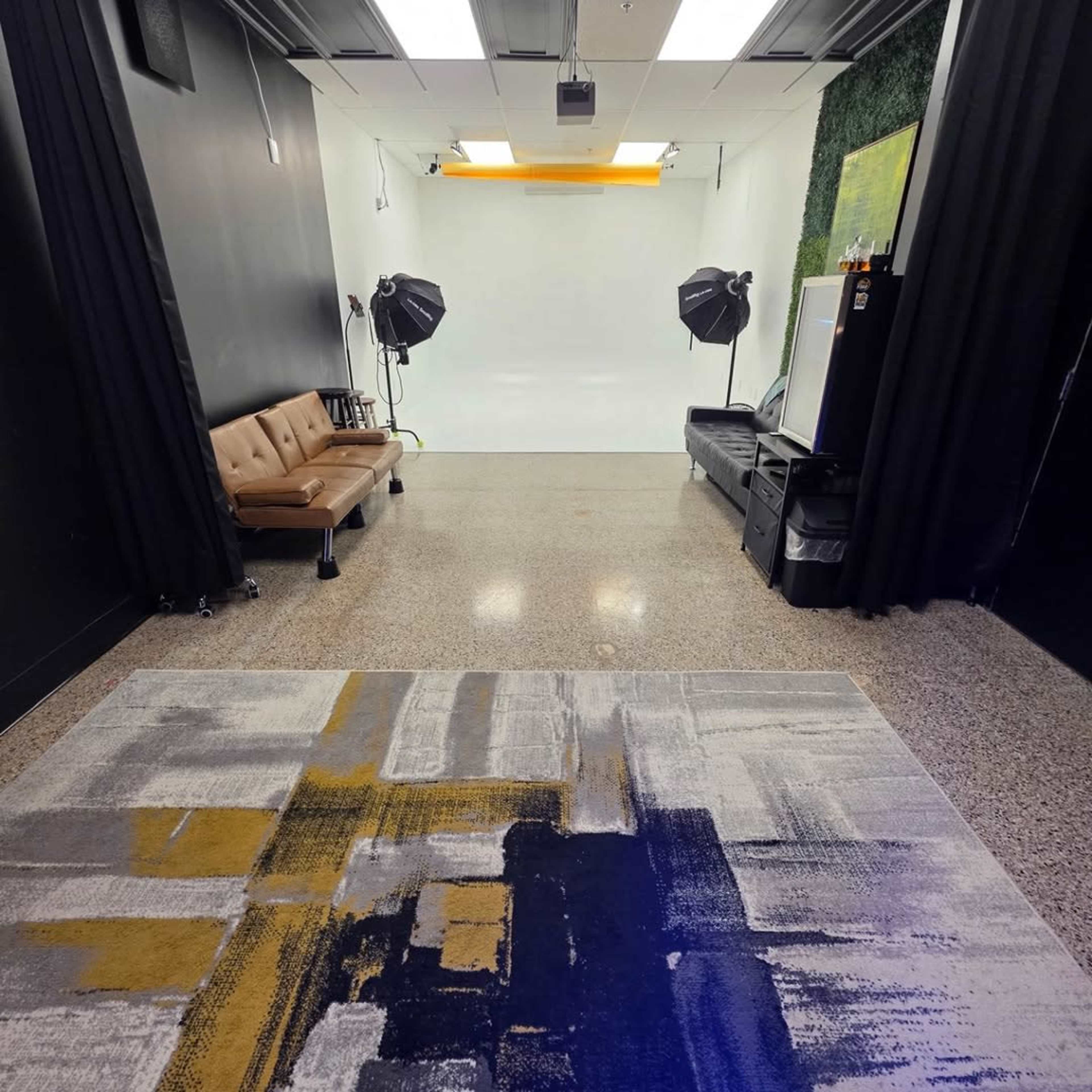 The image shows a photography studio with a light-colored backdrop, two studio lights, a brown sofa, and a patterned rug on a polished floor.